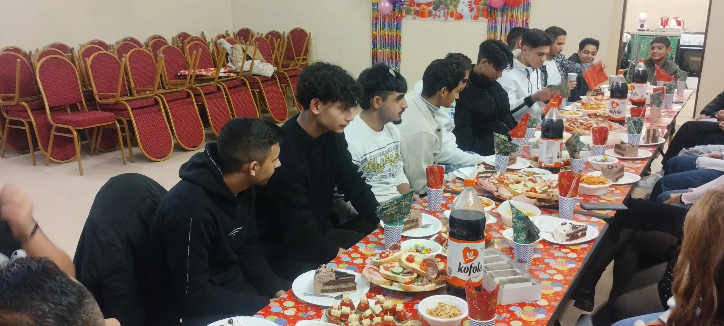 Group of young men sitting at a long table with festive food and drinks, celebrating indoors with Christmas decorations.