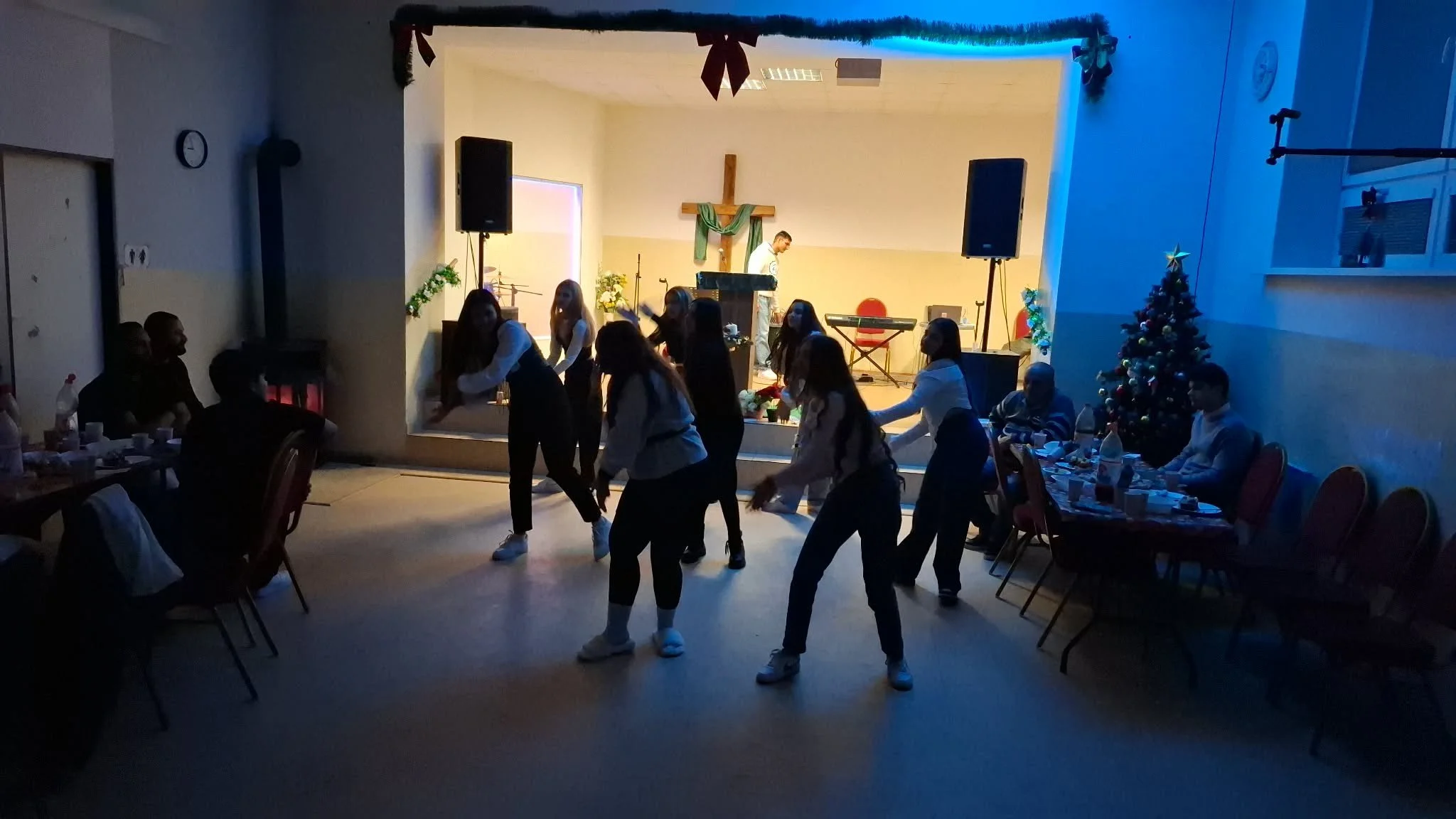 People dancing in a church hall decorated for Christmas, with a Christmas tree to the right and a stage at the back featuring a cross and musical instruments.