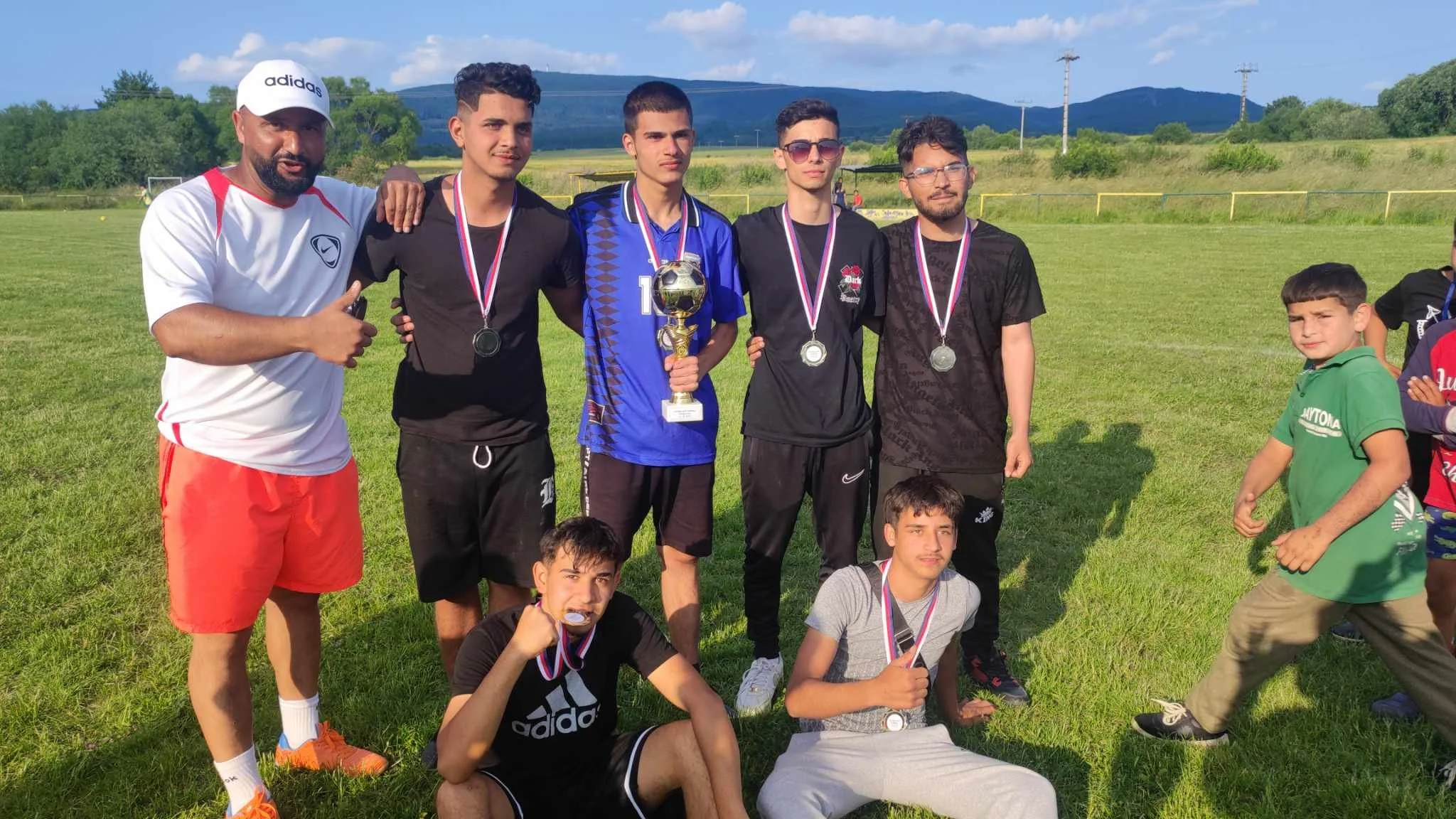 A group of young men and boys on a grassy field, celebrating with medals and a trophy after a soccer game or tournament. Five stand in the back, with two seated in front. They are outdoors with mountains and power lines in the background.