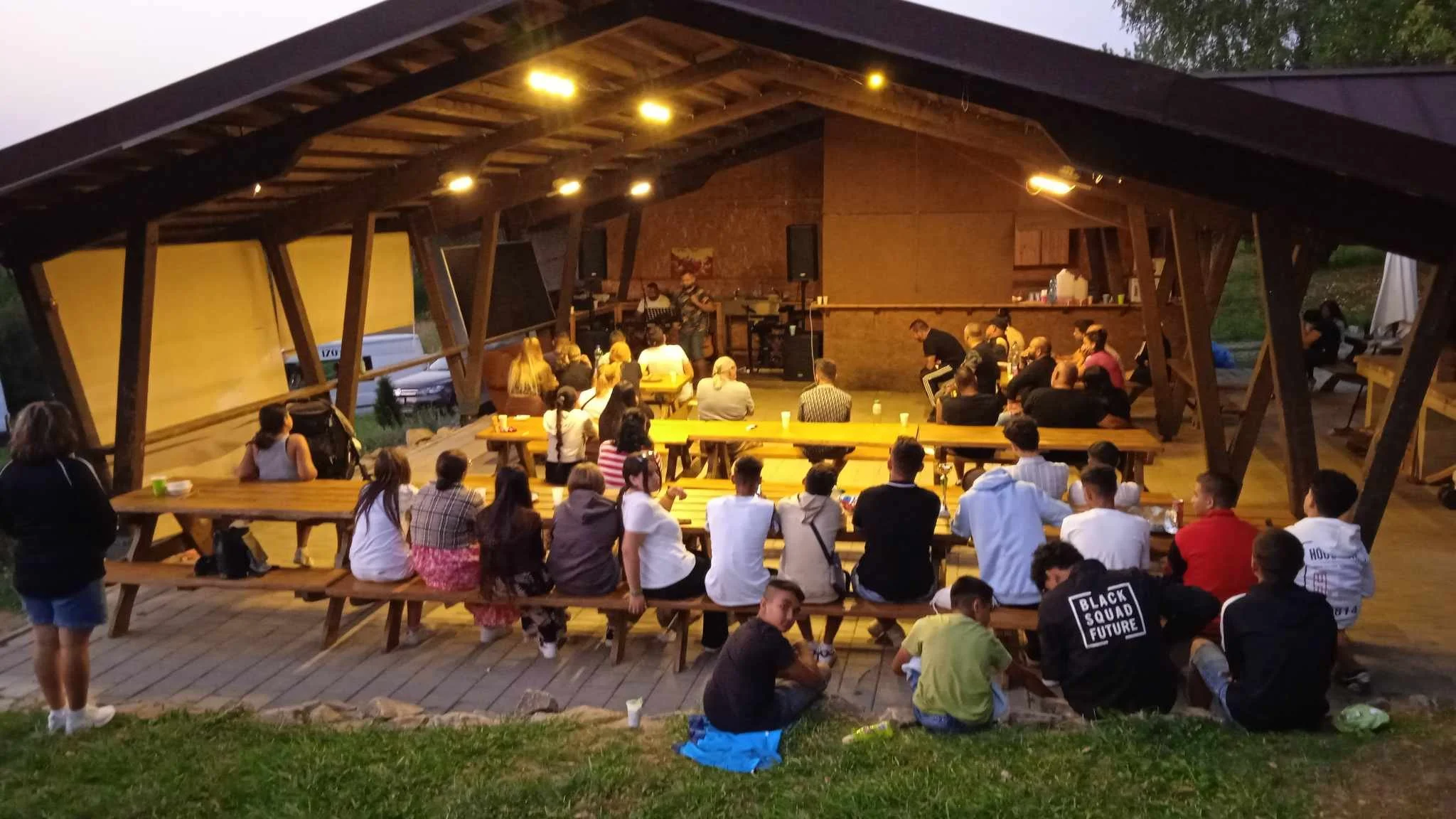 A crowd of people seated and standing at outdoor wooden benches and tables under a pavilion watching a live band perform.