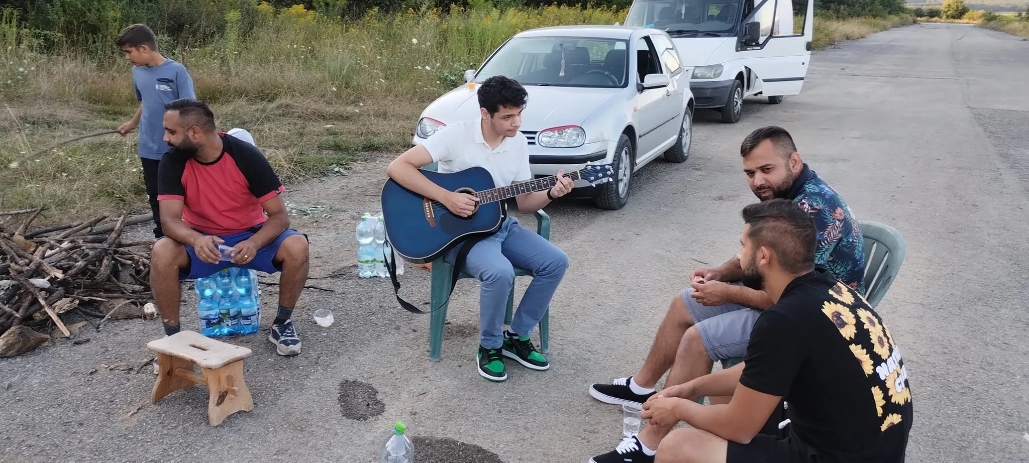 A group of people outdoors on a dirt road, sitting around a small campfire. One young man is playing a guitar while others are sitting and chatting. There are two cars parked behind them, a white one and a van, with a grassy field and trees in the ba