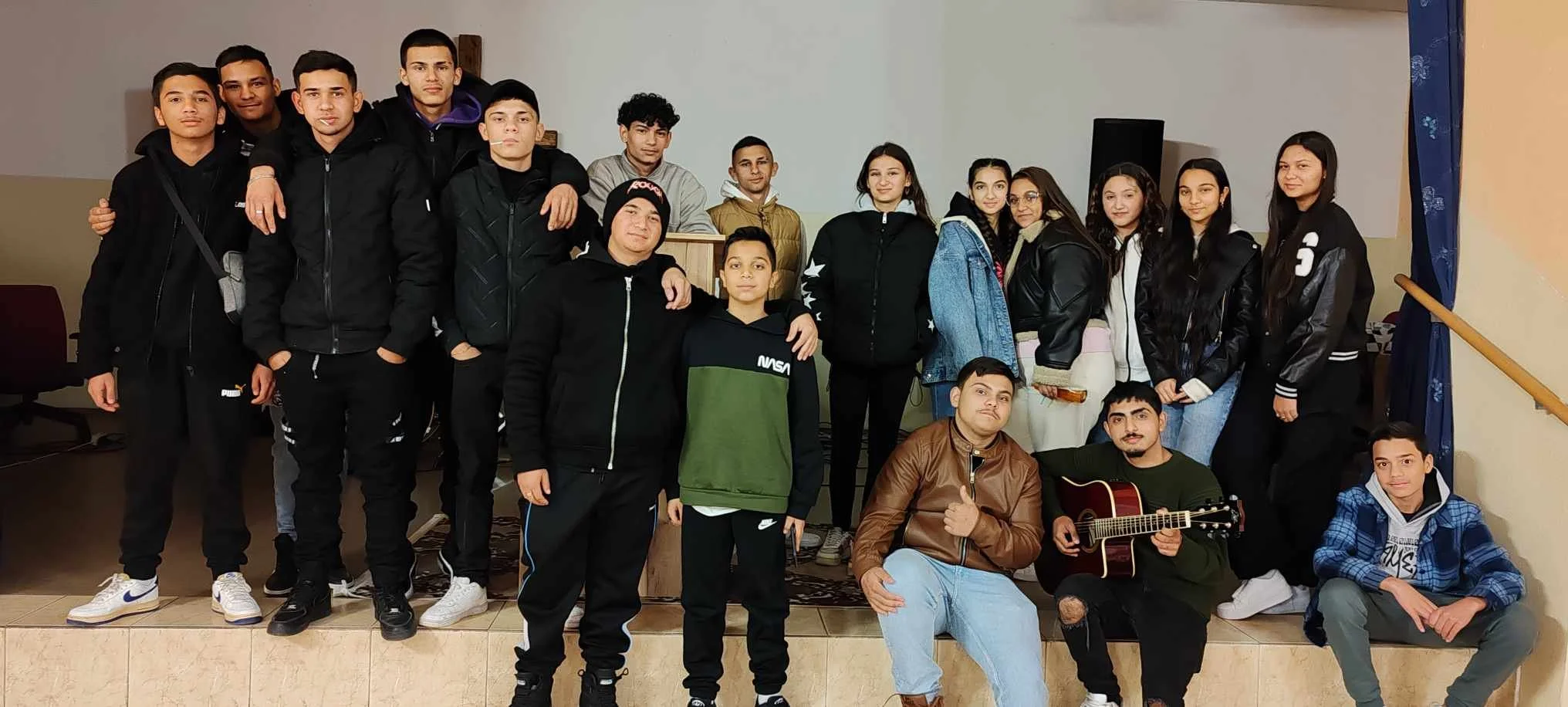 A large group of young people, mostly boys and girls, standing and sitting on stepped platform indoors. Some are dressed in casual jackets and pants, and two are holding a guitar. They appear to be in a social or school setting.