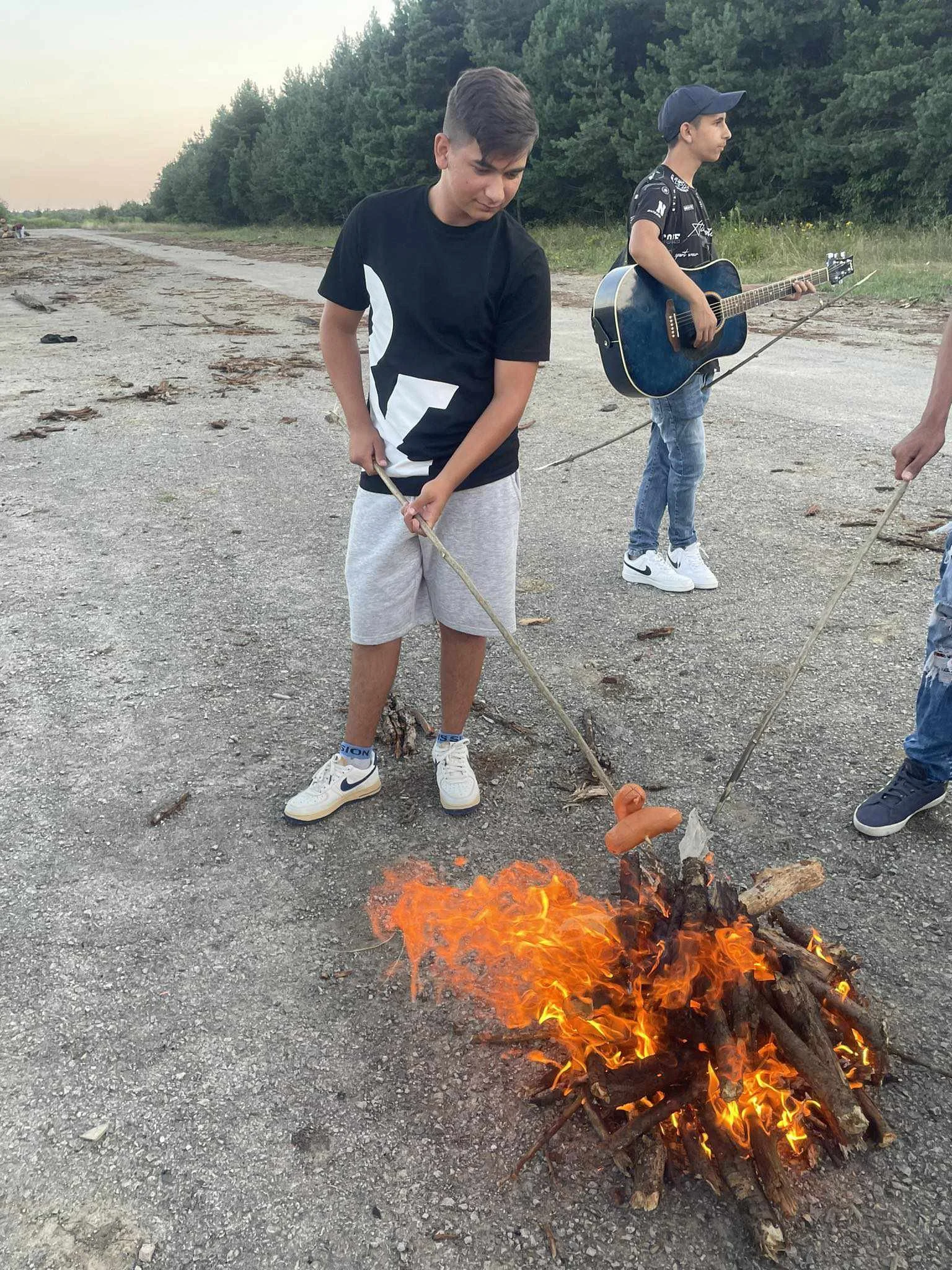Two boys roasting hot dogs over an outdoor campfire on a dirt path with trees in the background.