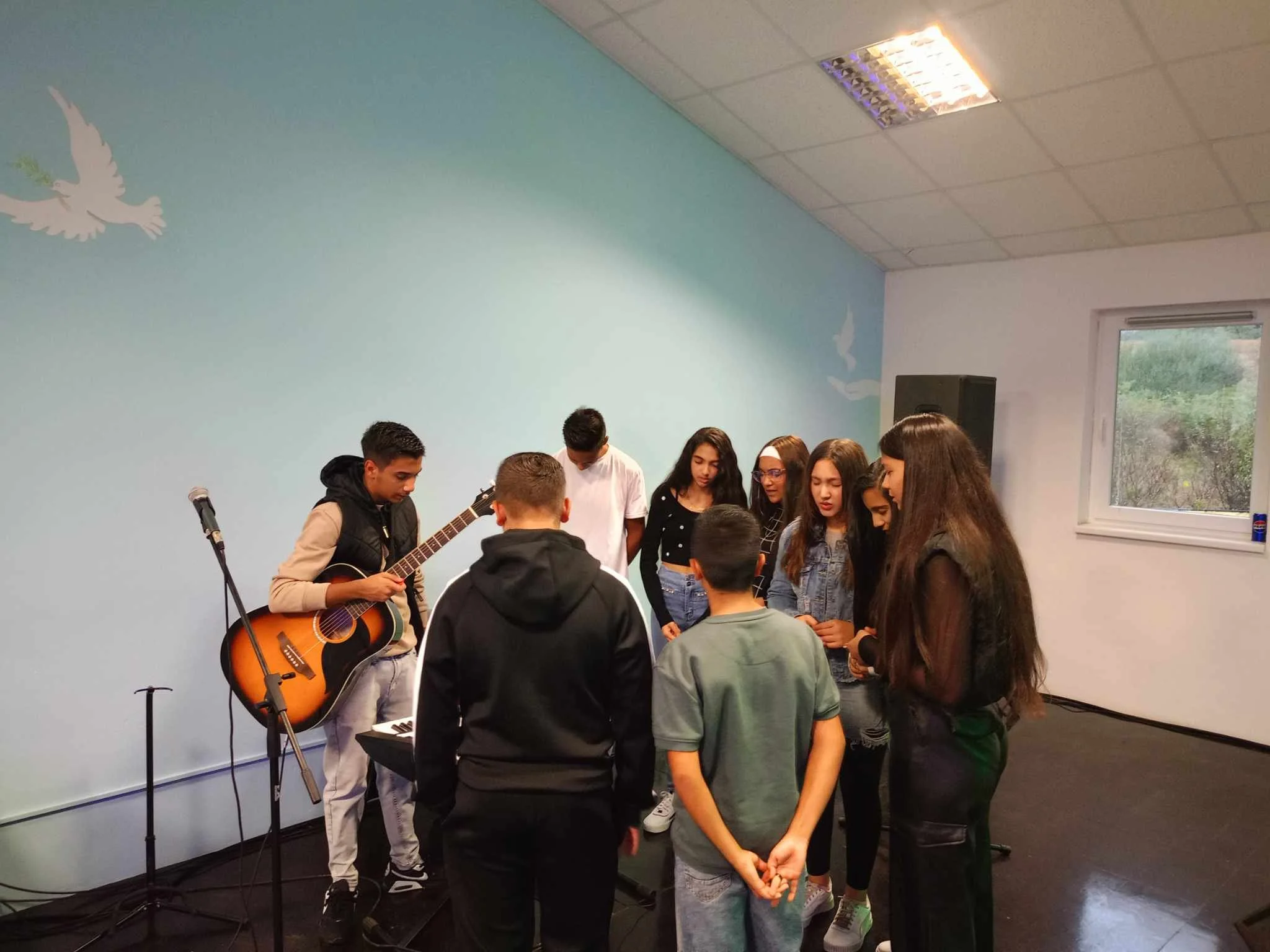 Group of young people huddled together listening to a guitarist playing an acoustic guitar in a room with light blue wall decorated with white doves.