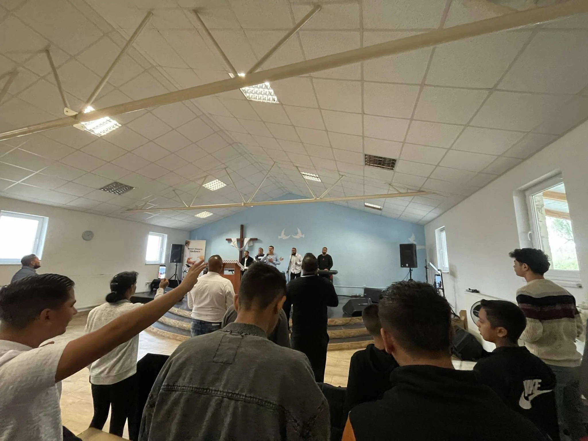 People gathered in a church or community hall during a service or event, with some standing and others seated, facing a stage with a cross and a band or choir performing.