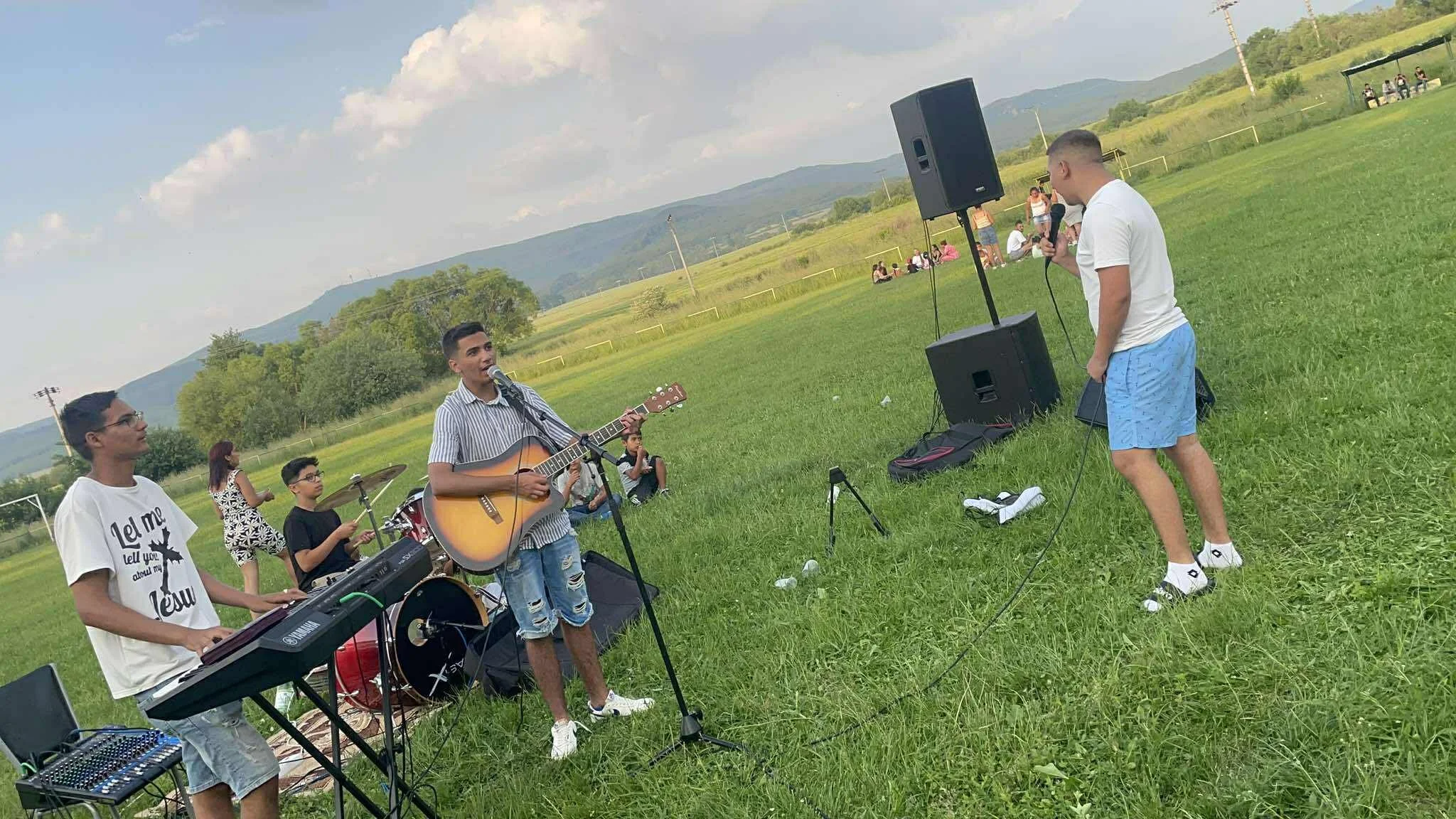 A group of young musicians performs outdoors on a grassy field with mountains in the background. One person sings and plays guitar, another plays keyboard, and a third is on drums. A person in shorts and a white t-shirt is speaking into a microphone,