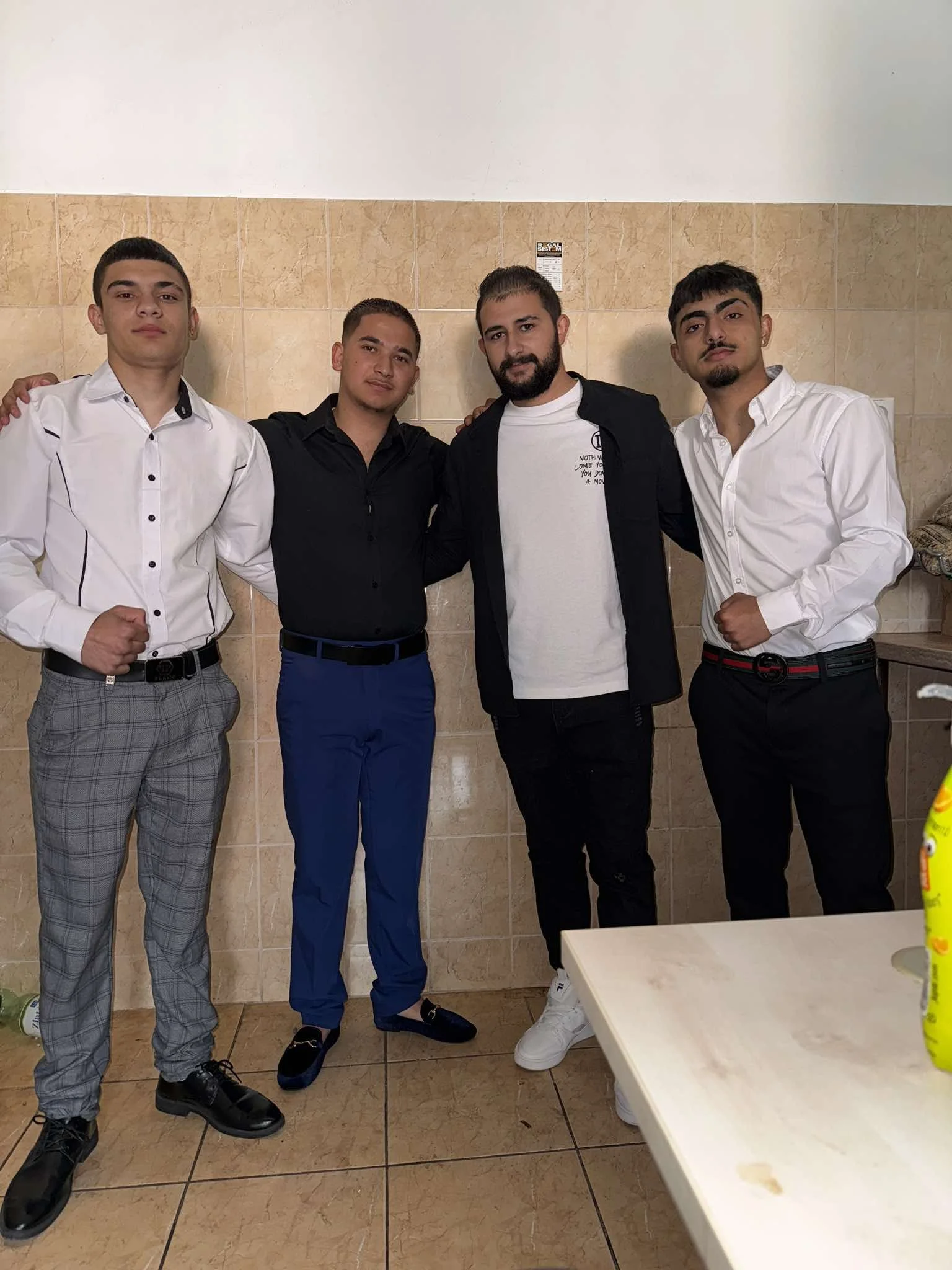 Four young men standing closely together against a beige tiled wall, with arms around each other's shoulders, dressed in formal and semi-formal attire.