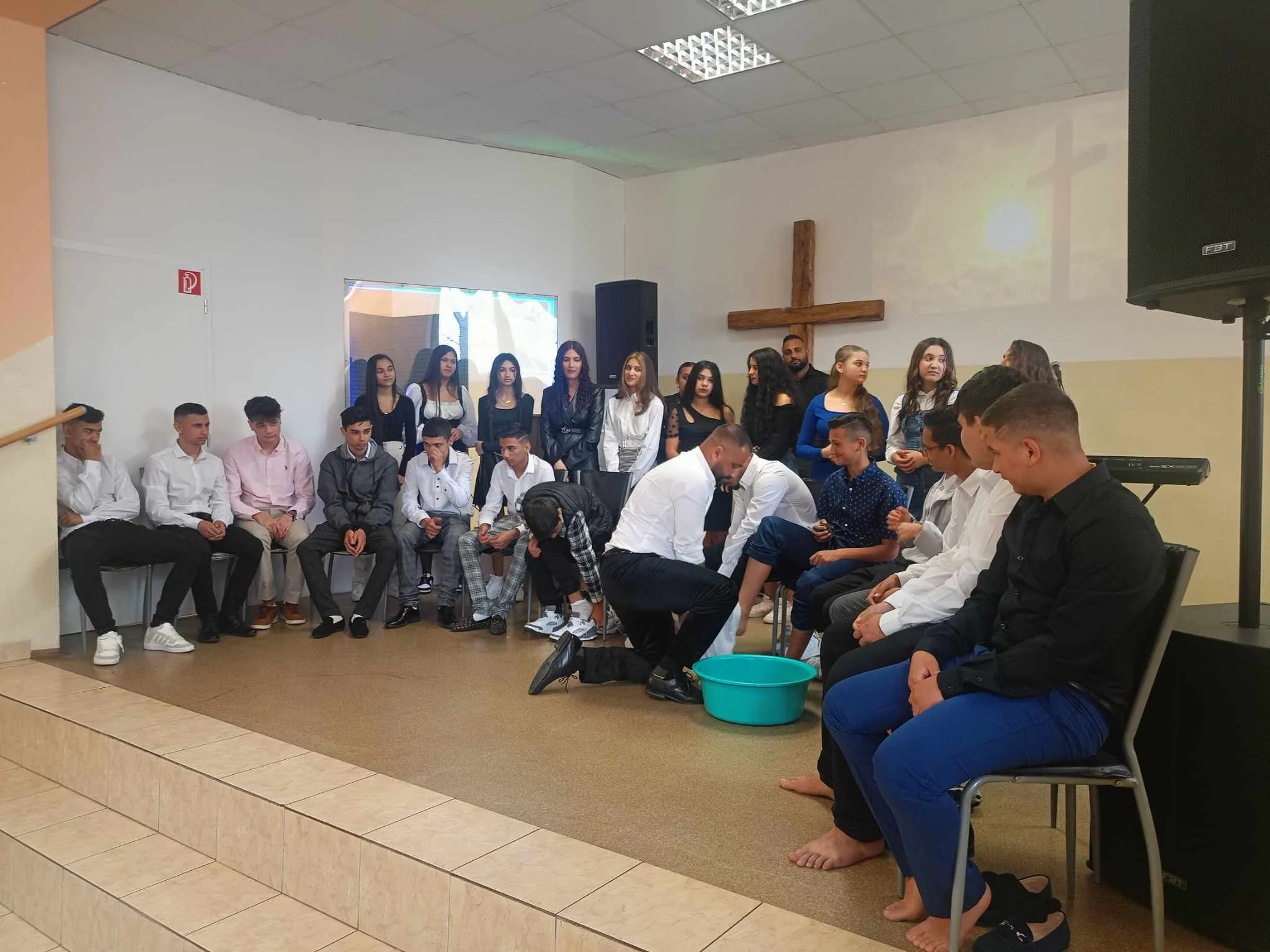 Group of young people gathered in a church or religious setting with a wooden cross on the wall, some sitting on chairs and others standing, participating in a baptism ceremony.