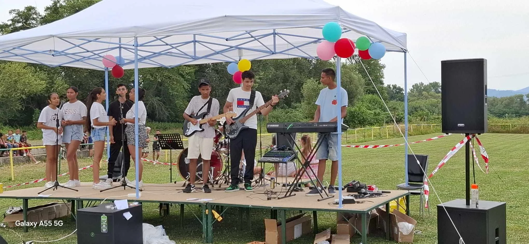 Group of young people performing music on an outdoor stage under a white canopy decorated with colorful balloons during a community event.
