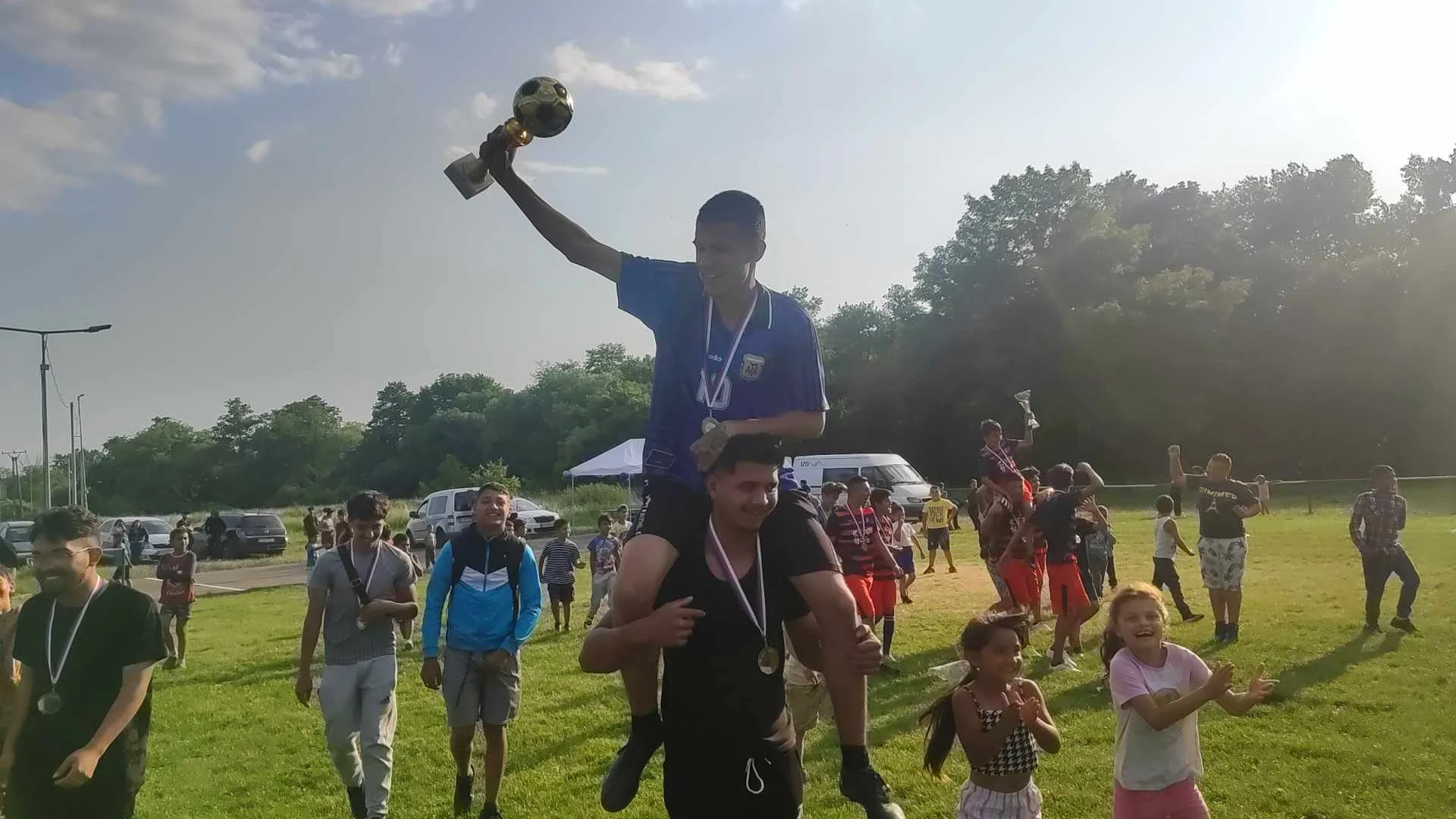 A young man celebrating a victory by sitting on someone’s shoulders, holding a trophy, with a crowd and children around him outdoors during daylight.