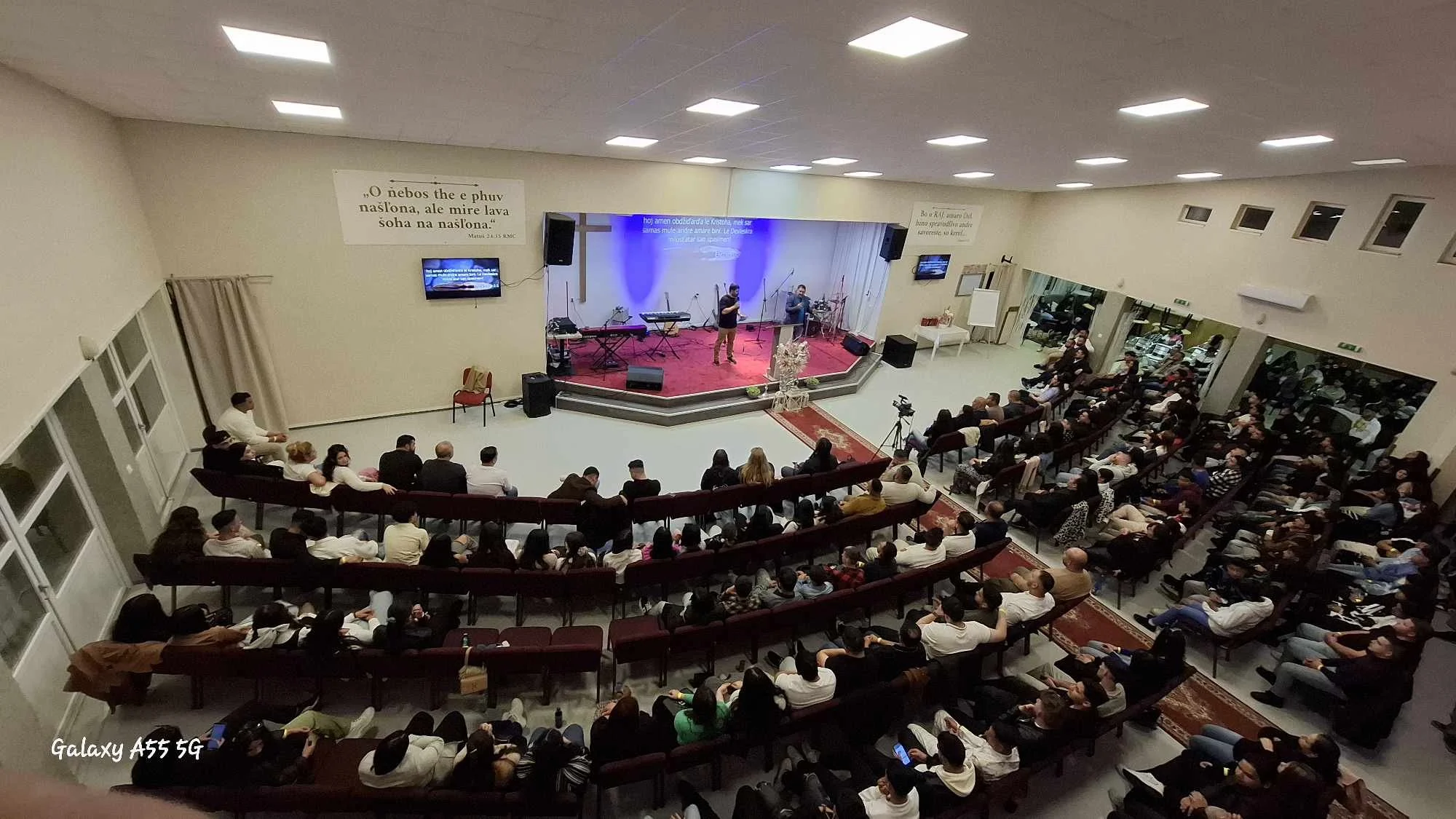 A church or auditorium with a stage where two speakers are addressing an audience. The room is filled with rows of chairs occupied by a diverse group of people. The stage has musical instruments and microphones, and there are screens displaying text.