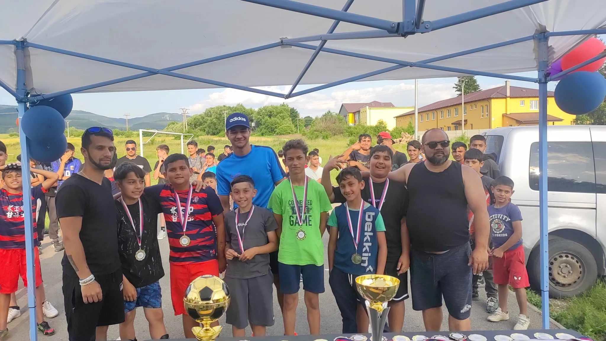 Group of young boys and adult men posing with medals and trophies at a sports event outdoors