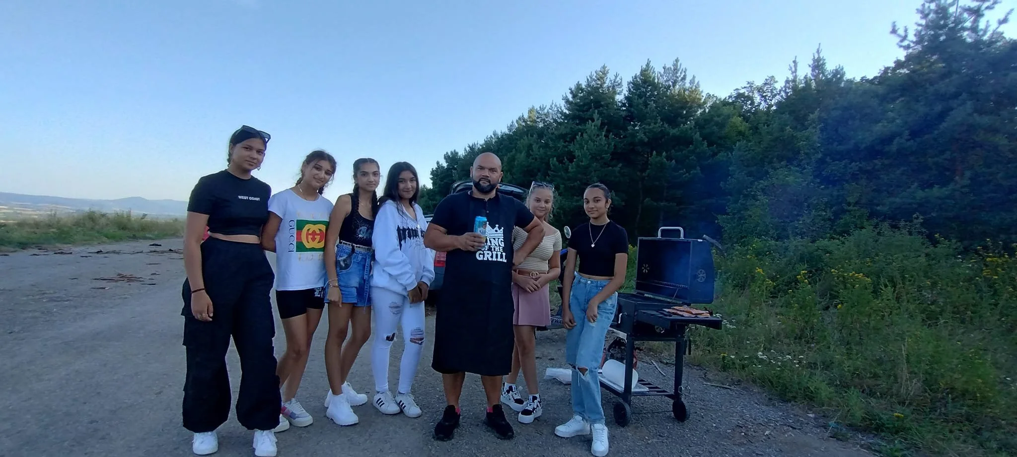 Group of six young women and one man standing outdoors near a barbecue grill, with trees in the background, during what appears to be evening or late afternoon.
