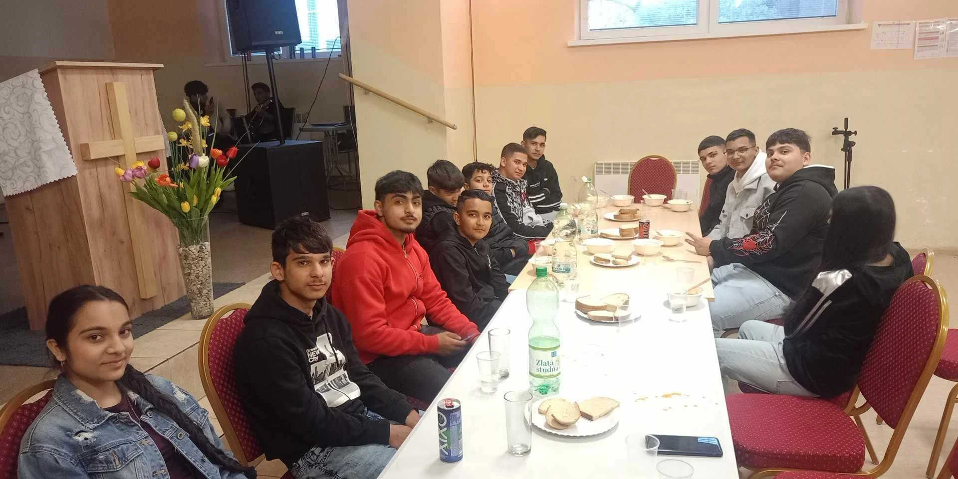 A group of young people sitting around a long table in a room, with some food, bottles, and cups on the table. There is a large flower arrangement and a wooden cross on a stand in the room. The group appears to be at a gathering or celebration.