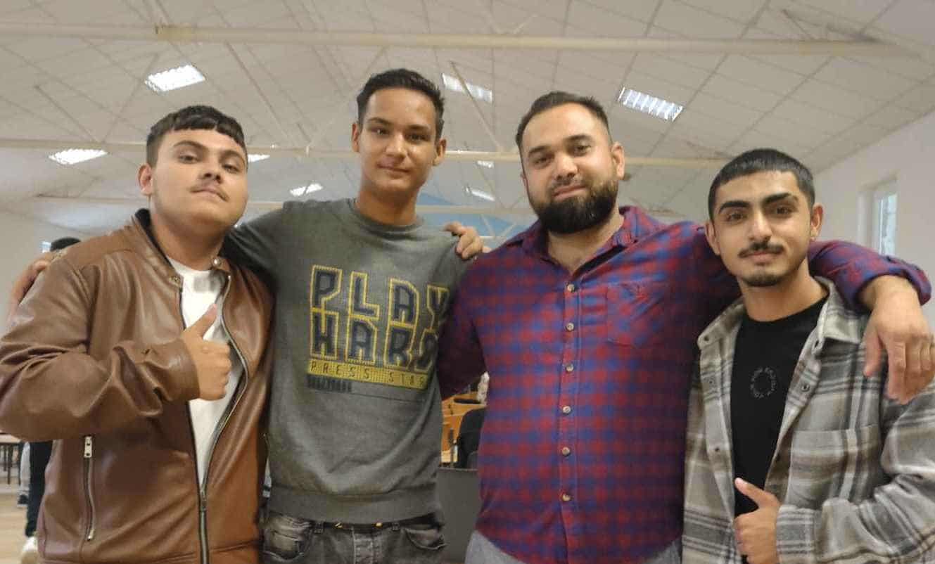 Four young men standing together indoors with their arms around each other, smiling at the camera.