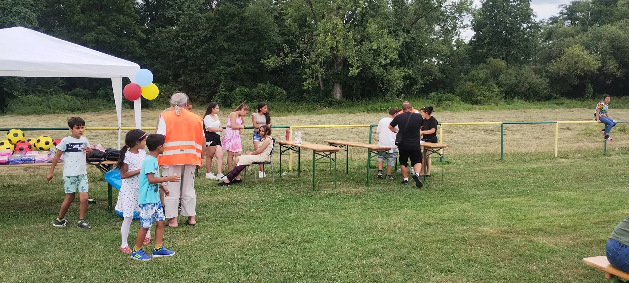 Children and adults gather outdoors at a park or field for a community event, with a white canopy tent decorated with balloons, tables with supplies, and a grassy area with trees in the background.