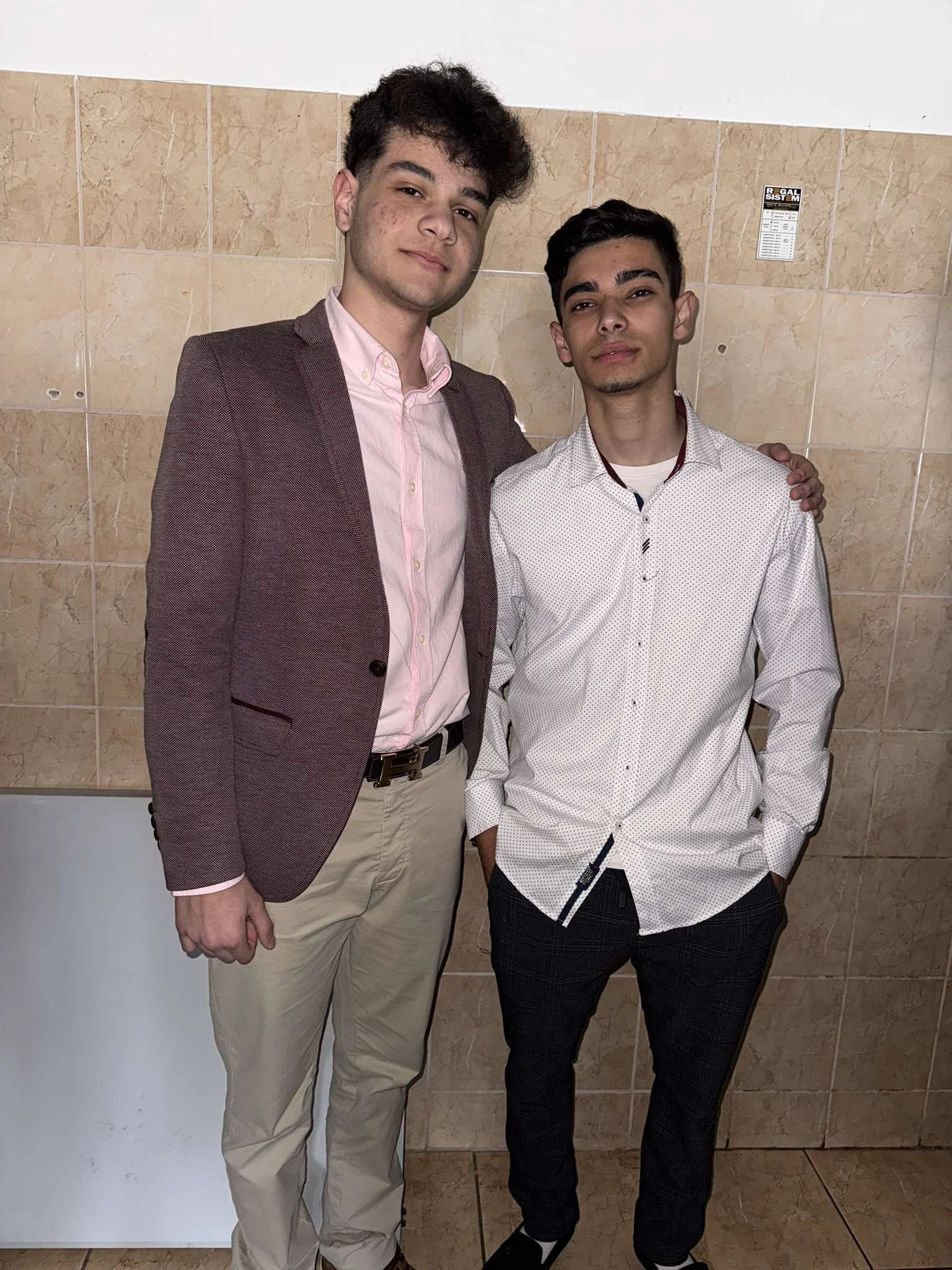 Two young men standing together against a tiled wall, one with a blazer and the other with a white shirt, posing for a photo.
