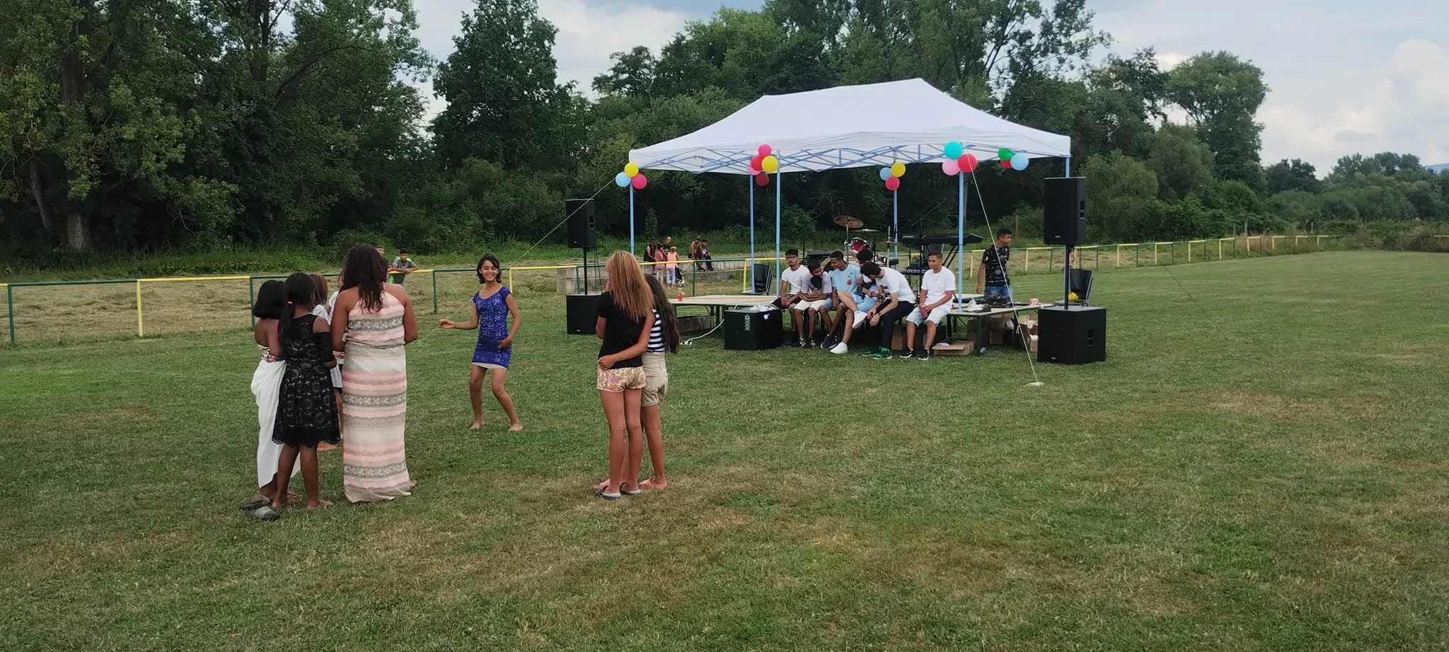 Group of children and teenagers at an outdoor event with a music stage, balloons, and grassy field, surrounded by trees.