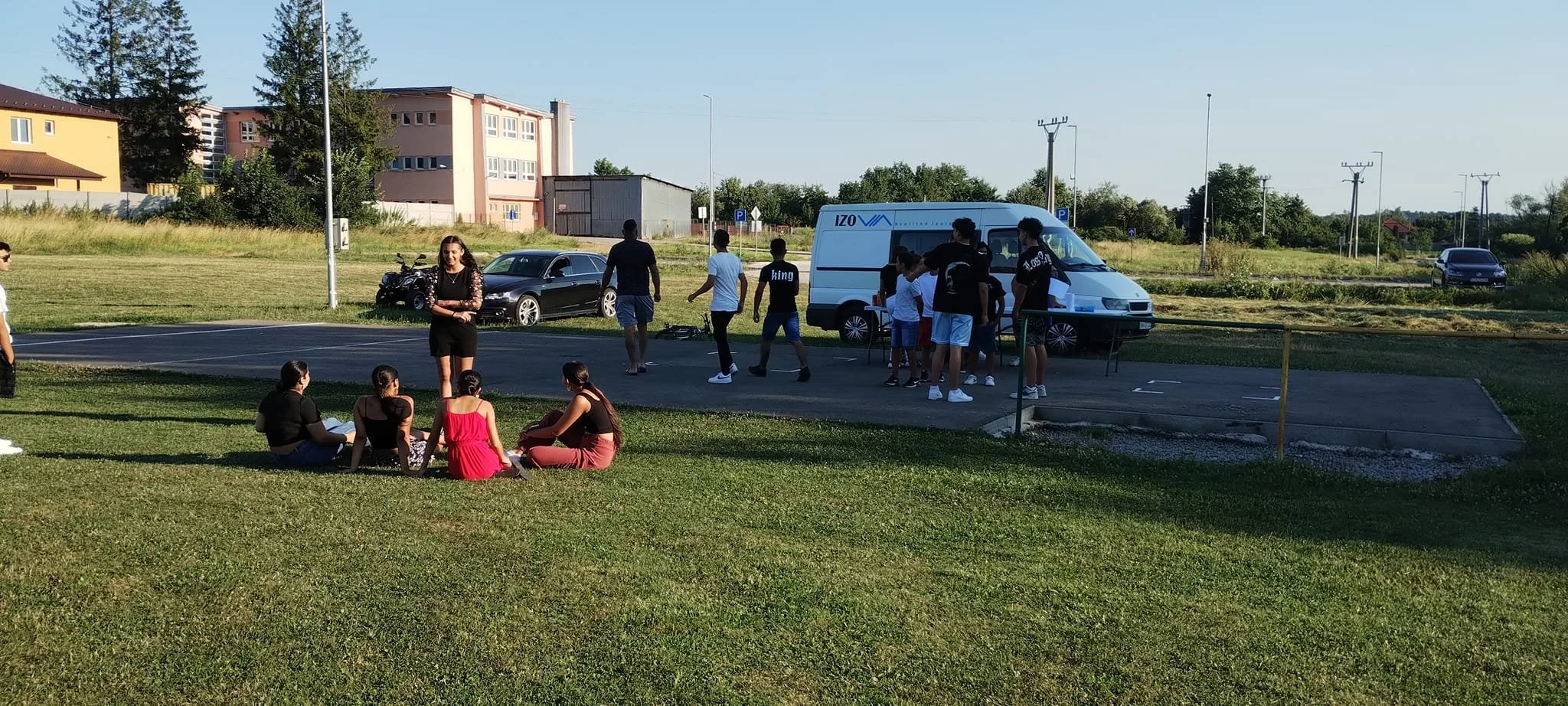 A group of people gathered outdoors near a white van and parked cars on a sunny day, with a grassy area and buildings in the background.