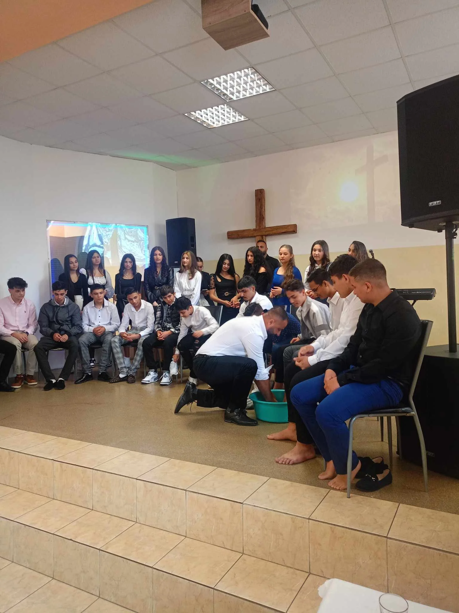 A group of people gathered in a church or meeting hall, with some sitting on chairs and others standing in the background. Two individuals in the foreground are kneeling and washing another person's feet, indicating a foot-washing ceremony.