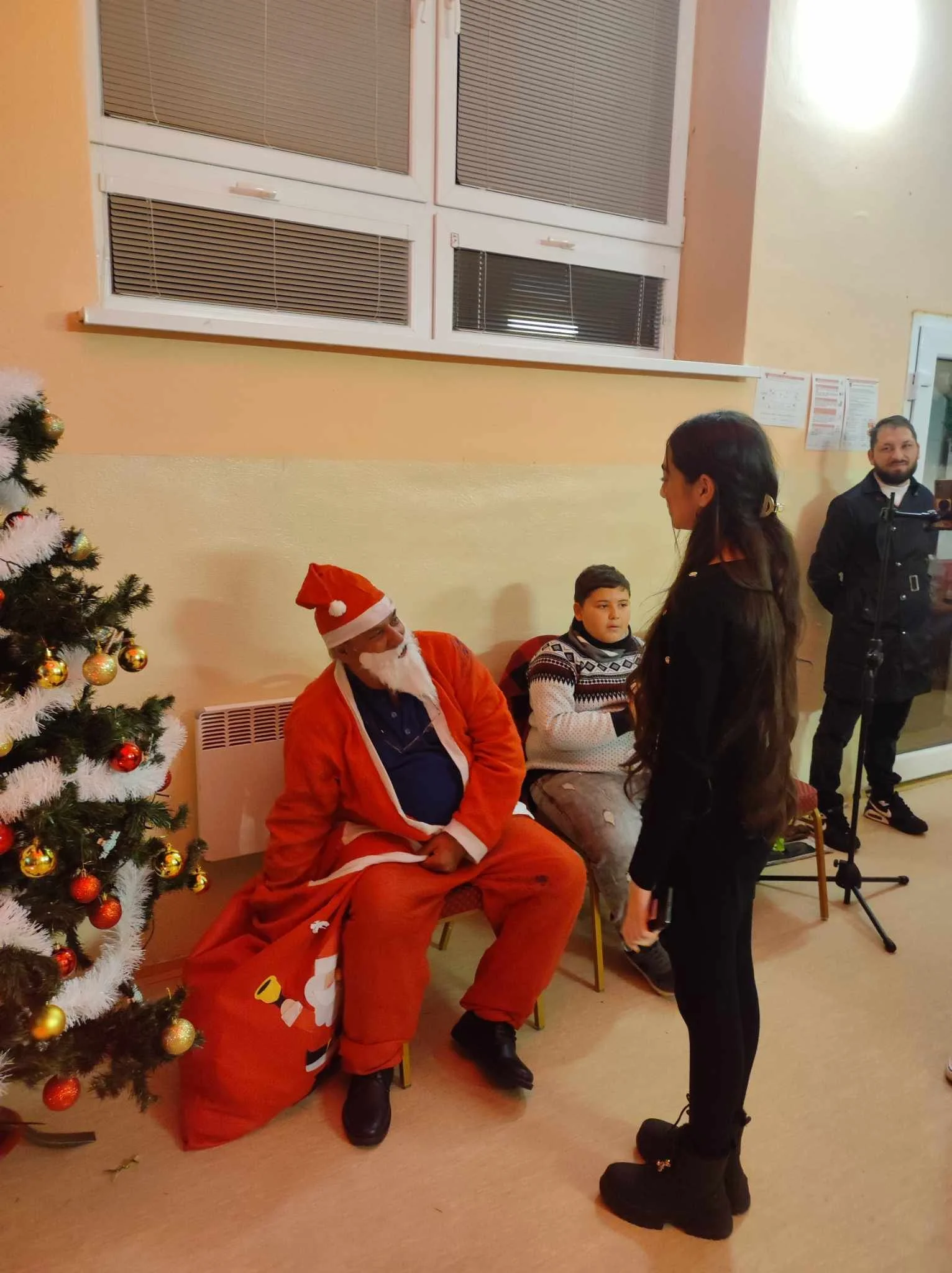 A girl standing in front of a man dressed as Santa Claus, sitting near a decorated Christmas tree. A boy sits beside Santa, and a man is recording the scene. The setting appears to be indoors, with holiday decorations.