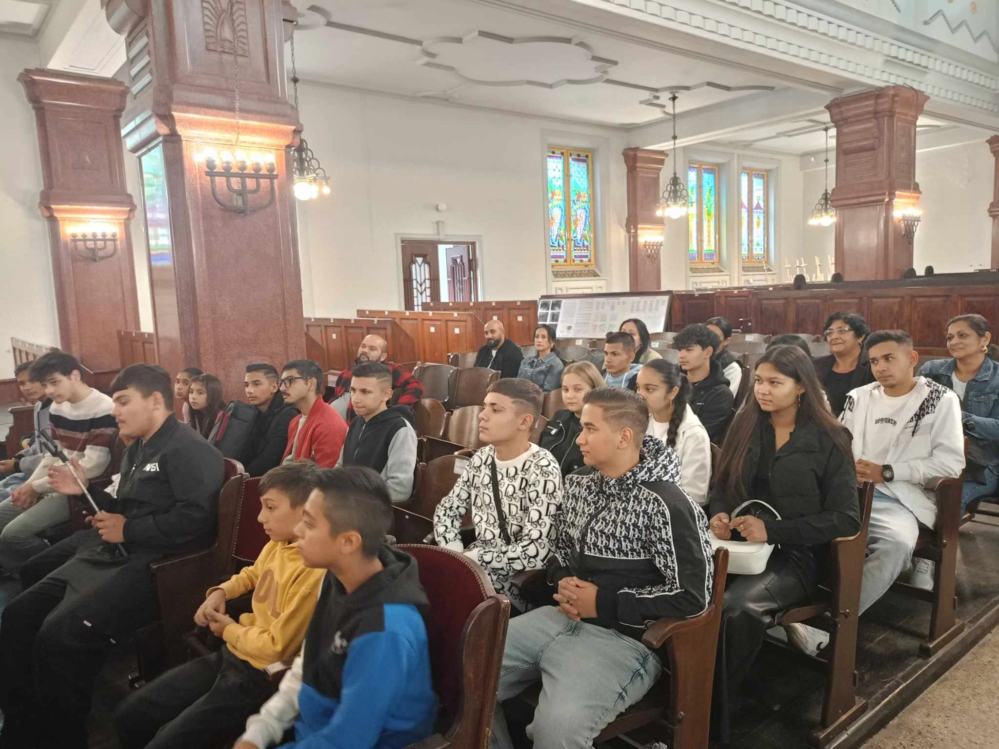 Group of people seated inside a church or cathedral, with stained glass windows and wooden pews.