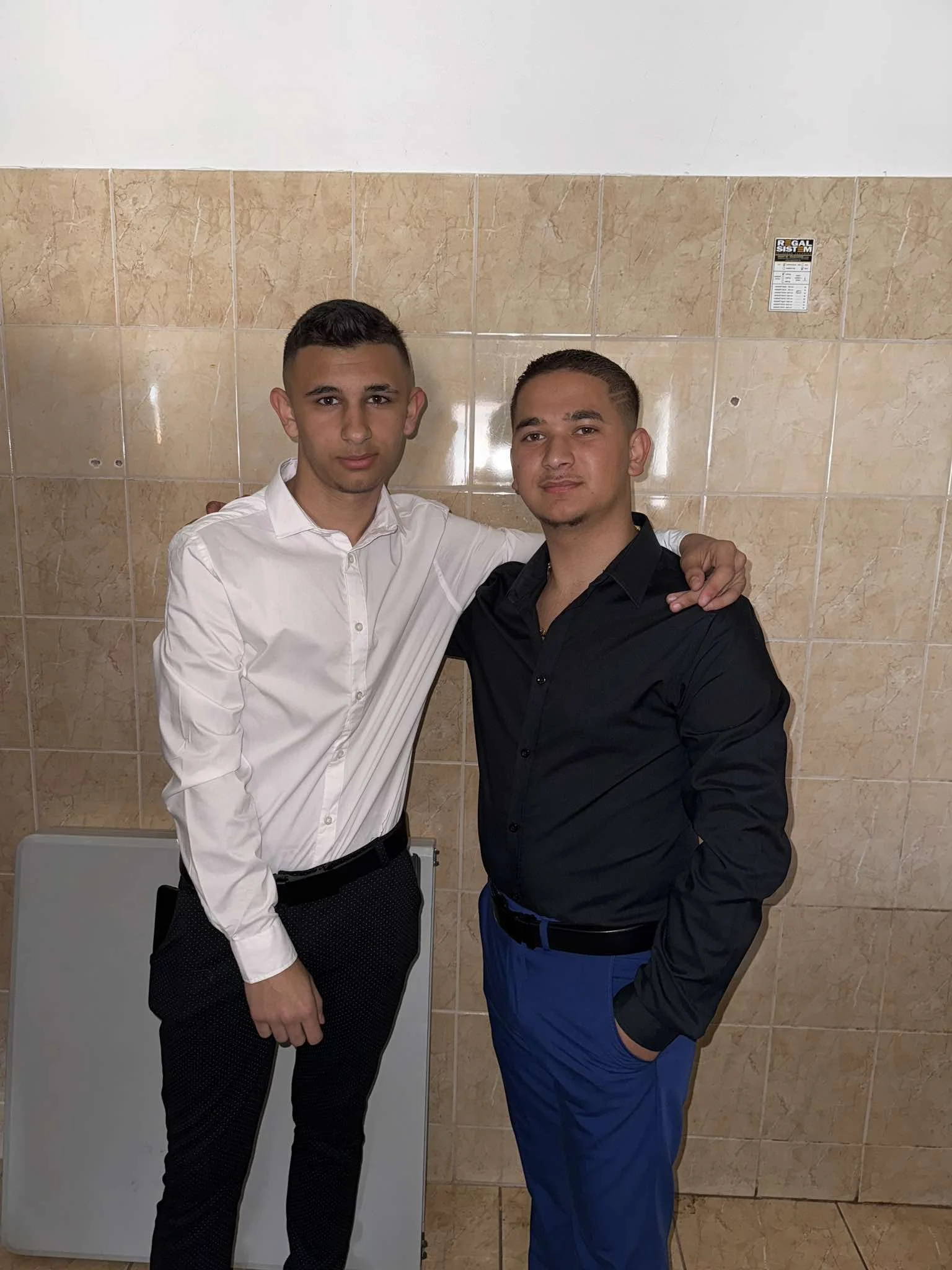 Two young men standing together indoors, wearing formal attire, with one in a white shirt and the other in a black shirt, against a beige tiled wall.