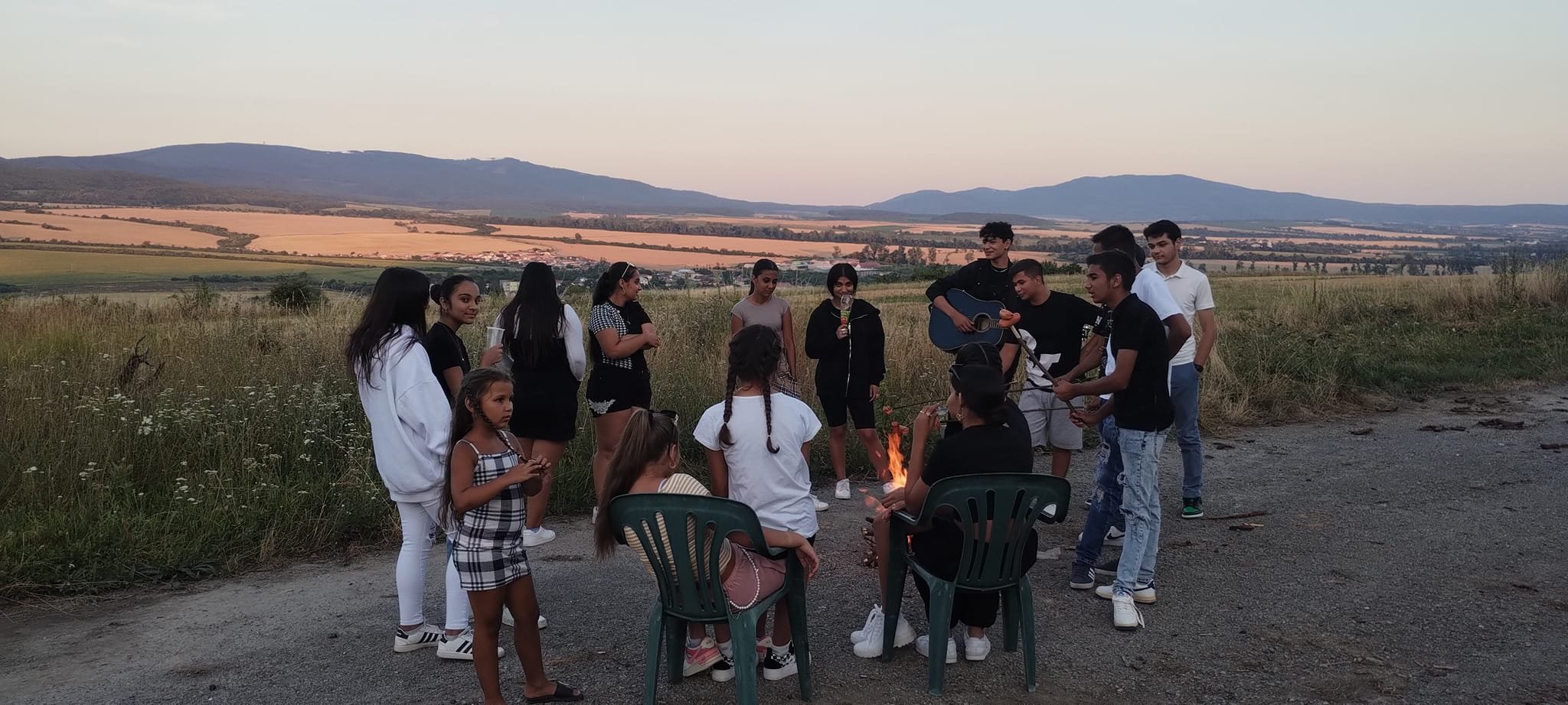 A group of young people gathered outdoors in a grassy field during sunset, with some sitting and others standing, playing and singing music around a small fire.