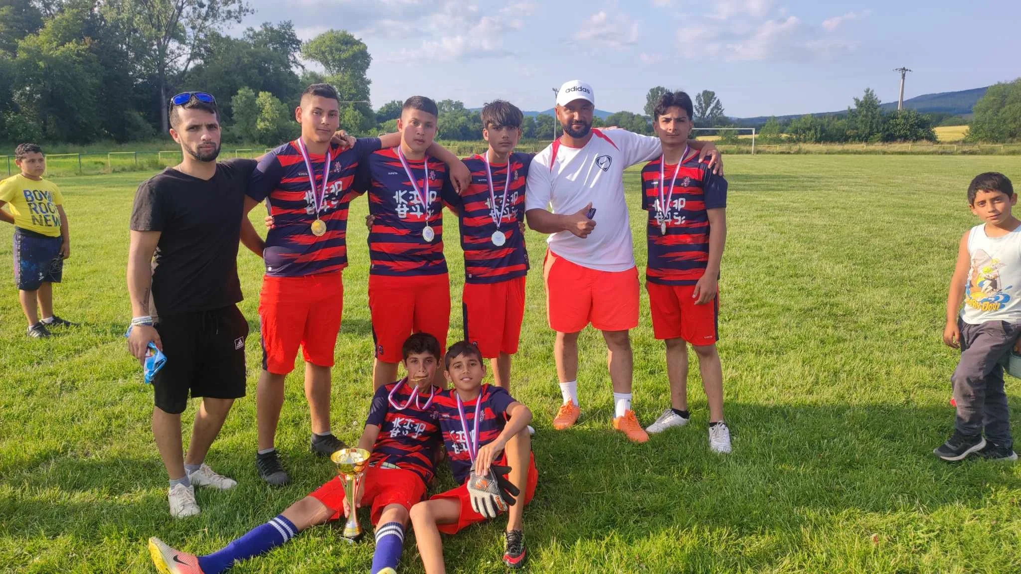 A group of young boys and their coaches celebrating on a soccer field with medals and a trophy, with a scenic background of trees and mountains.