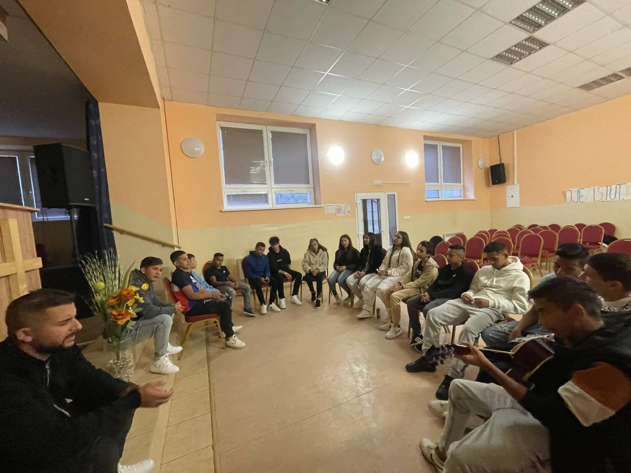 Group of students sitting in a circle in a classroom with orange walls and red chairs, some students are playing guitar, a teacher or facilitator is sitting with a flower arrangement nearby.