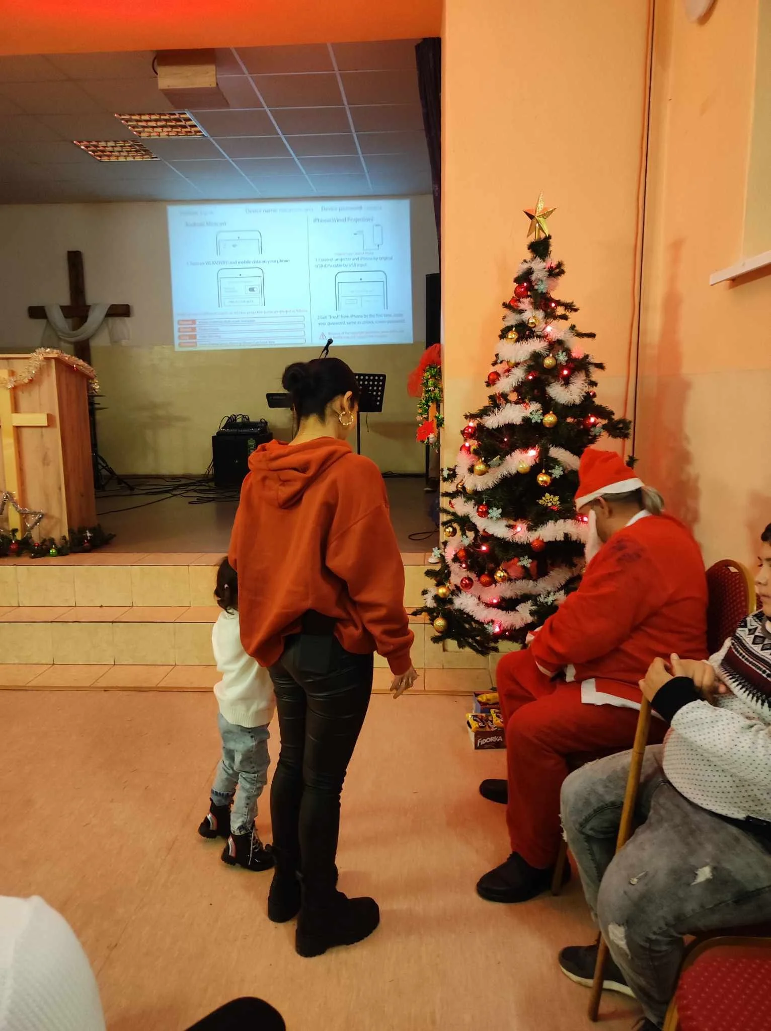 A Christmas scene in a community hall with a decorated Christmas tree, a person dressed as Santa Claus, and a woman and child standing near the tree. There is a stage with a projector screen in the background, and a cross on the wall.