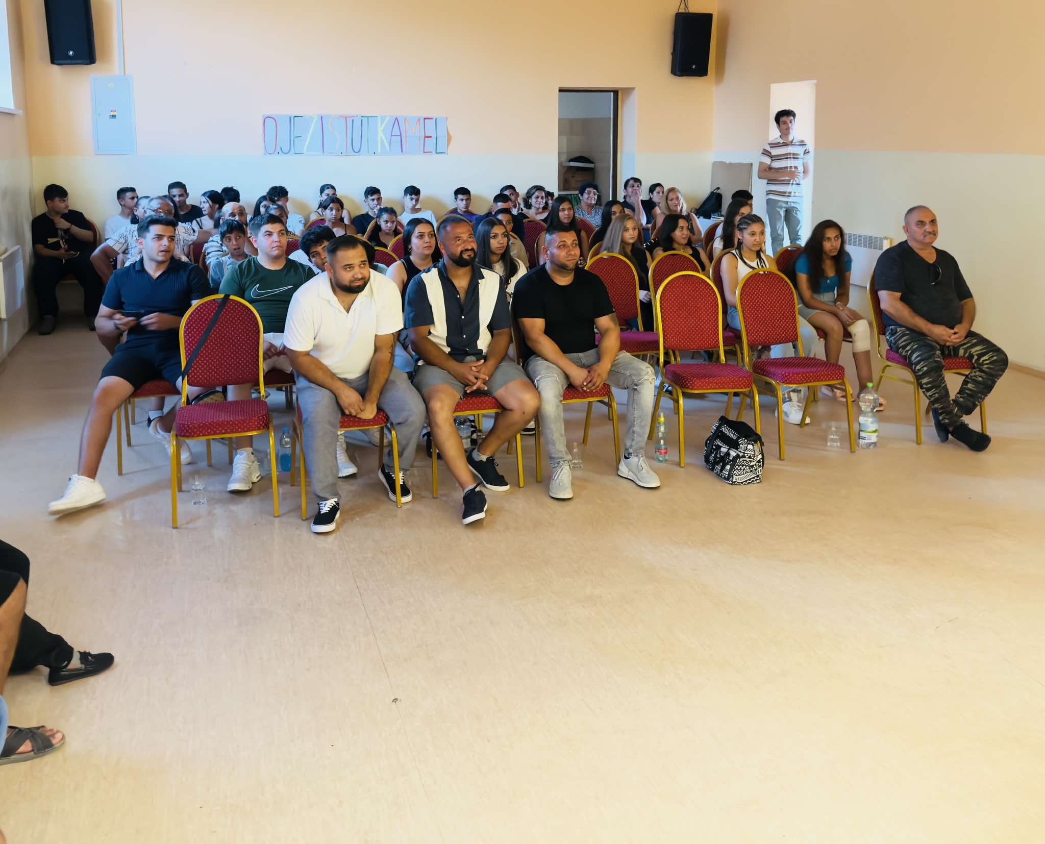 Group of people seated and standing in a room, attending an event or presentation, with a colorful banner on the wall.
