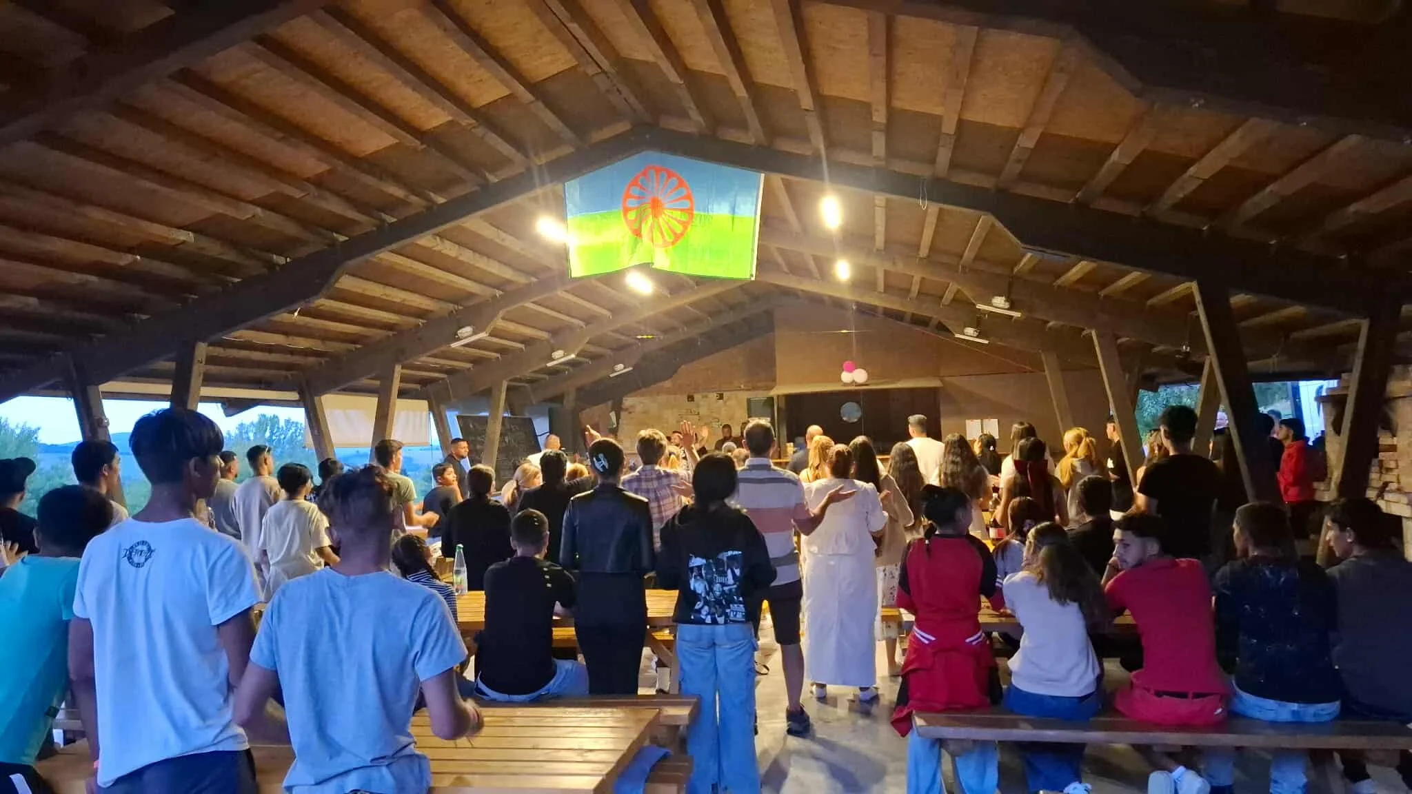 People gathered in a large indoor space with wooden beams on the ceiling, some standing and others sitting on wooden benches, attending an event or gathering with balloons and decorations.