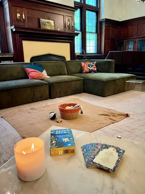 Living room with a green sectional sofa, wooden wall paneling, a window with blue-tinted glass, and a white marble table holding a lit candle, a deck of cards, and a book.