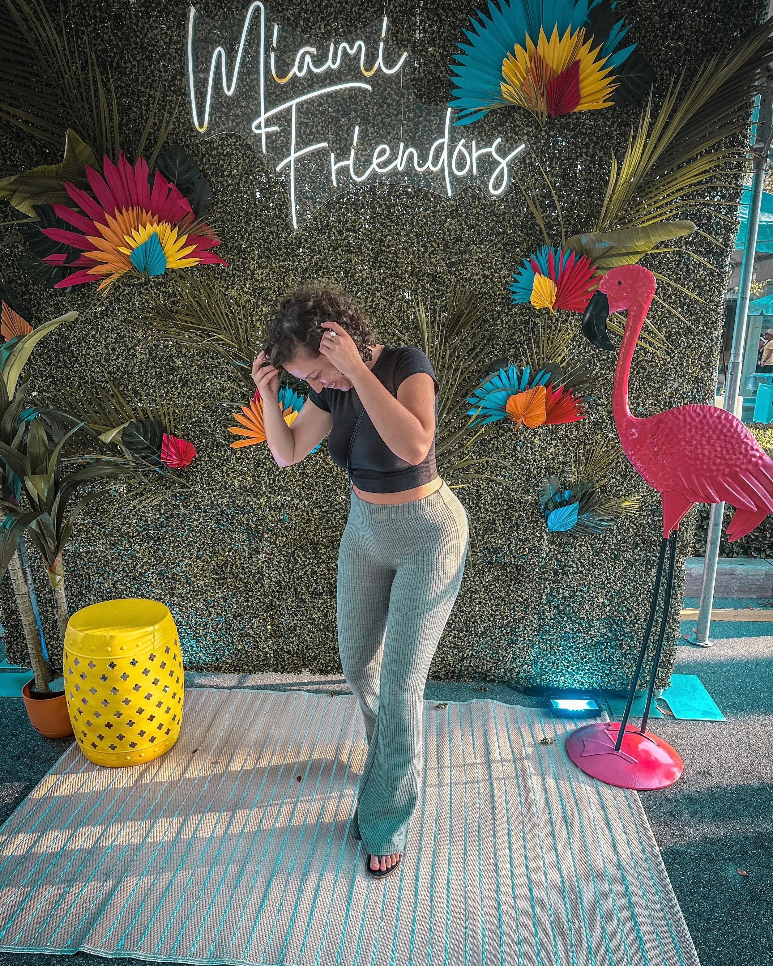 A woman with curly hair wearing a black crop top and beige pants standing in front of a tropical-themed backdrop with the neon sign that says 'Miami Fiends.' She is holding her head and smiling, with artificial plants, colorful paper fans, and a pink flamingo statue on her sides.