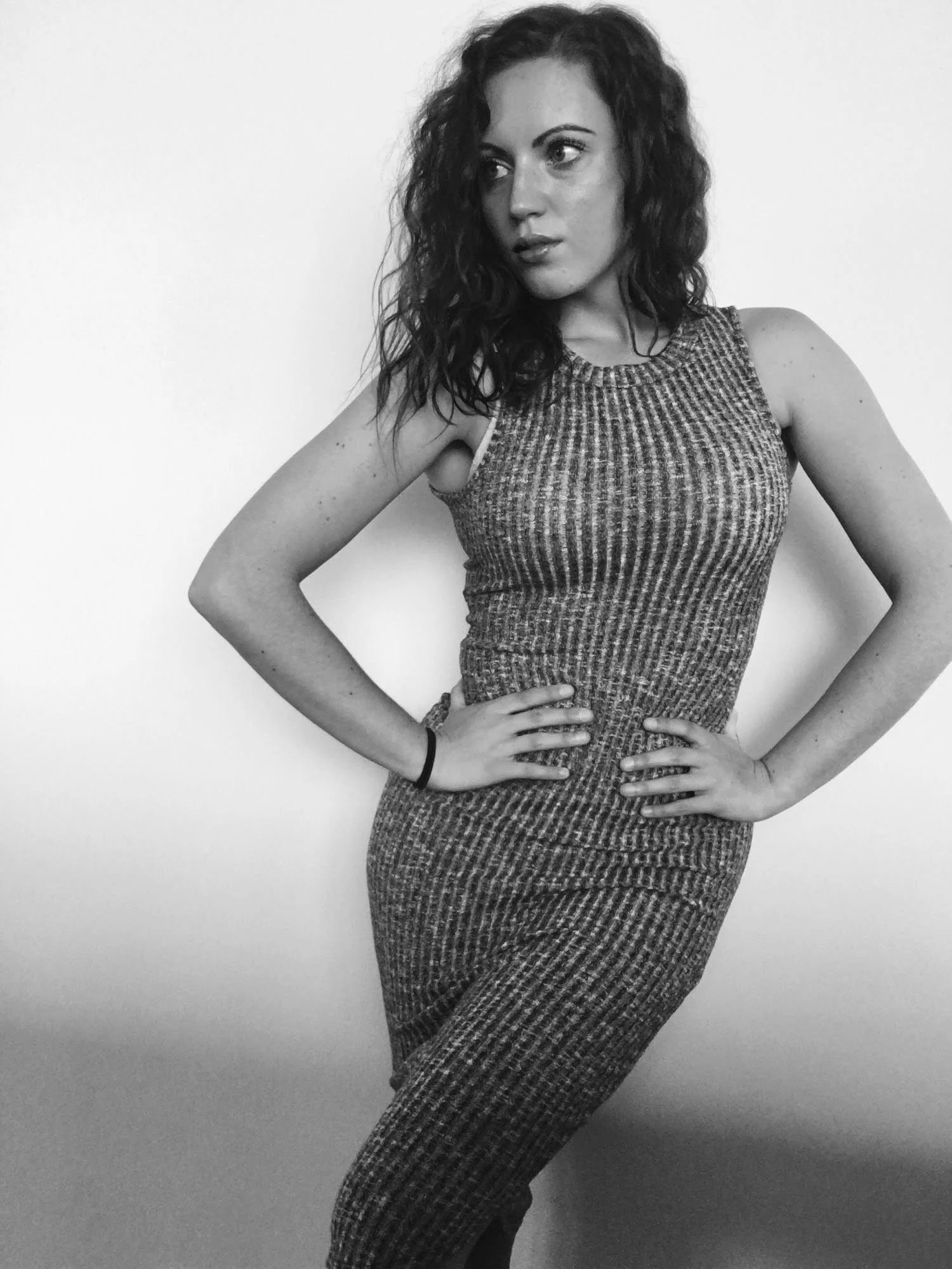 Black and white photo of a woman with curly hair standing against a plain wall, wearing a sleeveless patterned dress with her hands on her hips.