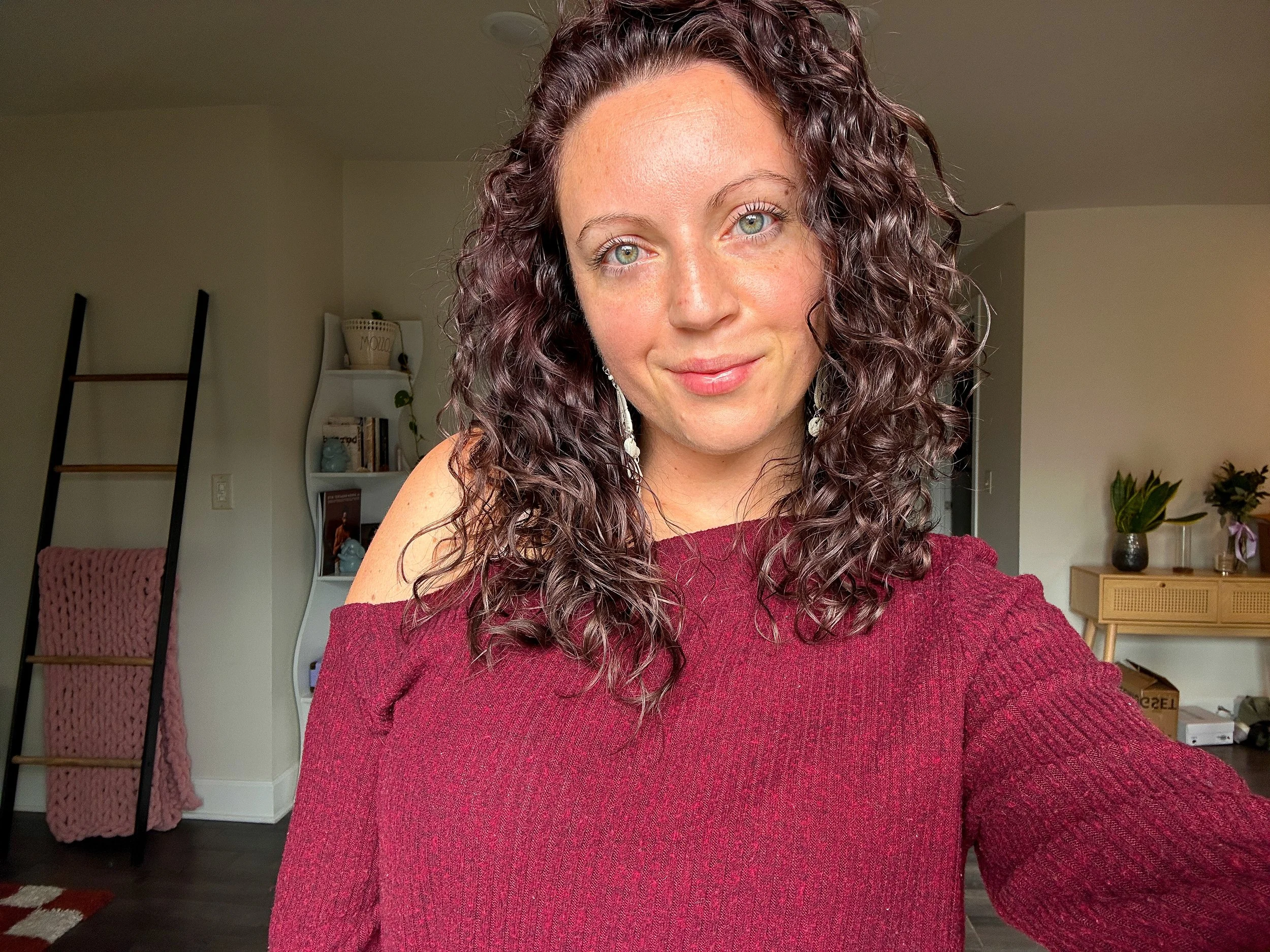 A woman with curly brown hair and blue eyes wearing a red cold-shoulder sweater and earrings, smiling at the camera in a cozy room with a bookshelf, plants, and decorative items in the background.
