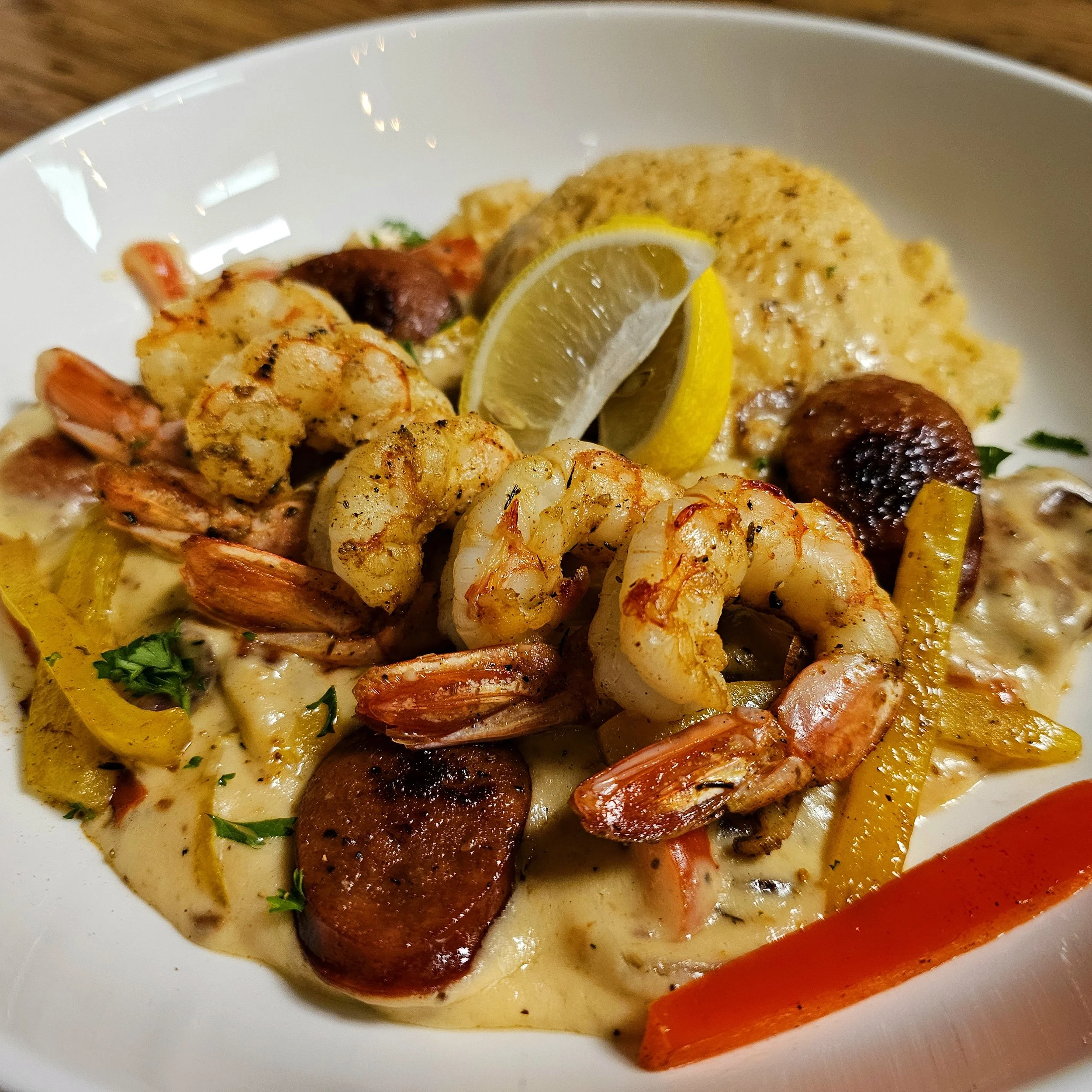 Seafood dish with shrimp, sausage, lemon wedge, mashed potatoes, and vegetables on a white plate.