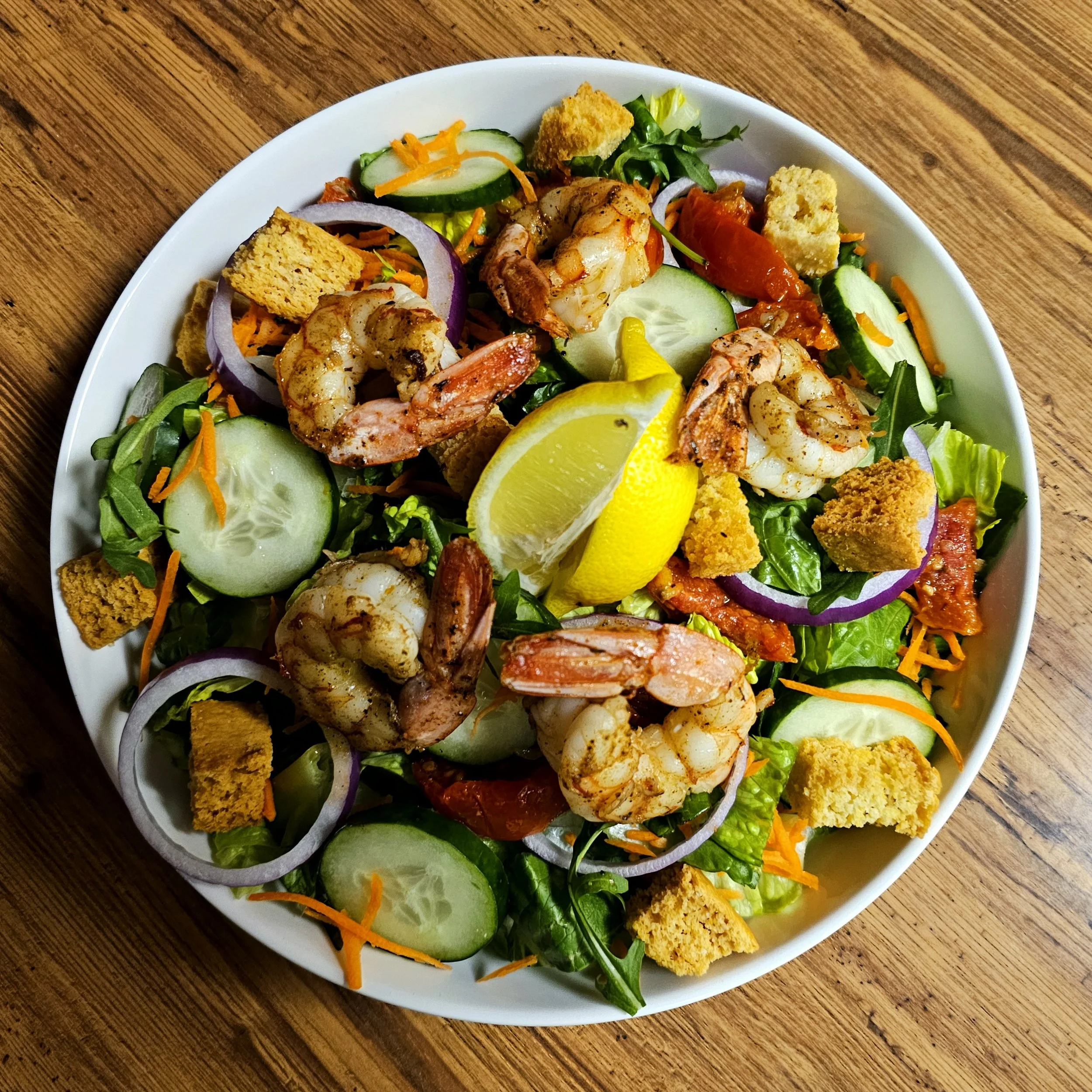 Seafood salad with shrimp, lemon wedges, cucumbers, red onions, shredded carrots, croutons, and mixed greens in a white bowl on a wooden table.