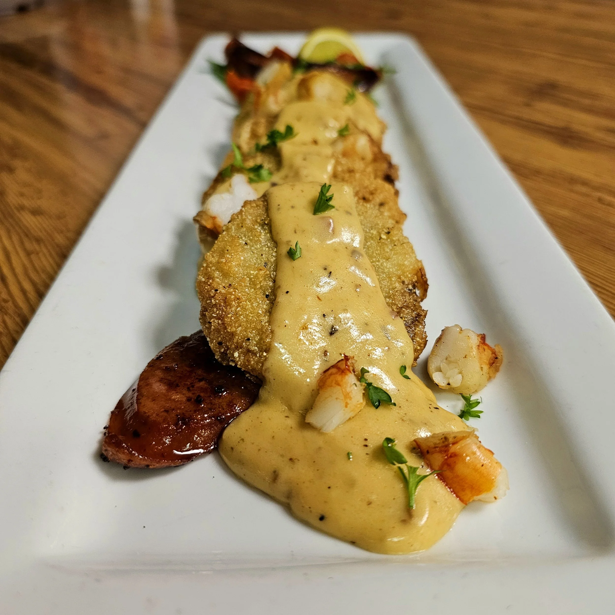 A white rectangular plate with fried fish fillet topped with creamy yellow gravy, garnished with parsley, accompanied by fried shrimp, sausage, and vegetables on a wooden table.