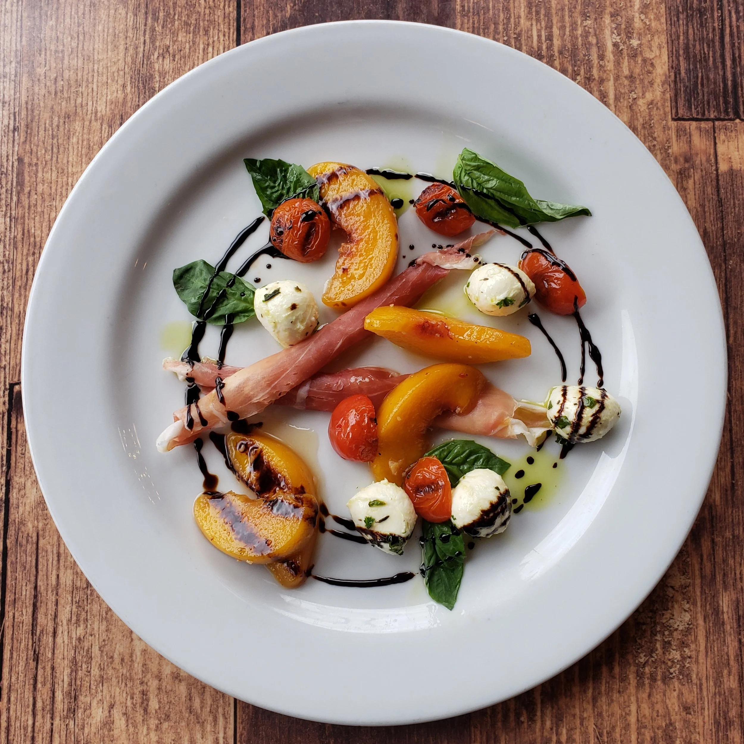 A plated appetizer with cherry tomatoes, fresh basil leaves, mozzarella balls, yellow peppers, prosciutto, and drizzles of balsamic glaze and olive oil on a white plate.