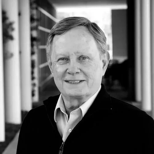 A black-and-white portrait of an elderly man smiling, wearing a collared shirt and a dark jacket, standing outdoors near a modern building with large windows.