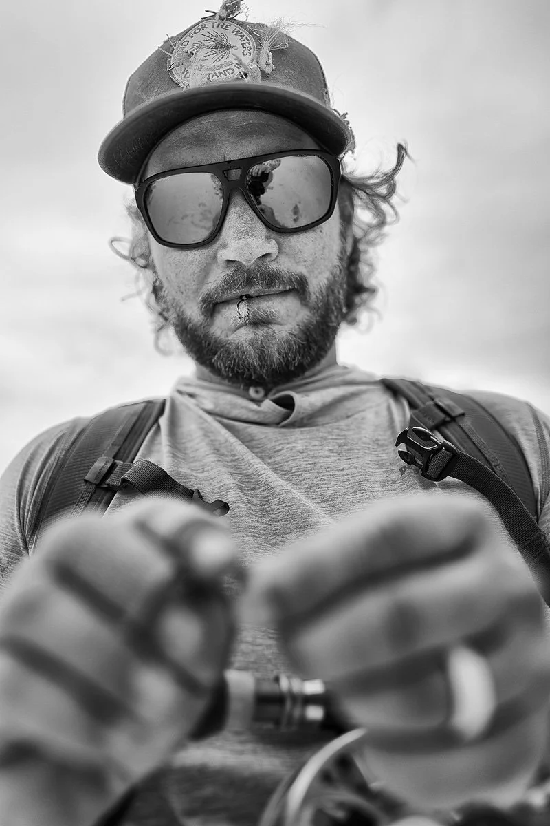 Drew Chicone, wearing a cap, large sunglasses, a gray t-shirt, and a backpack, looking down at fly he is tying onto his fishing line.