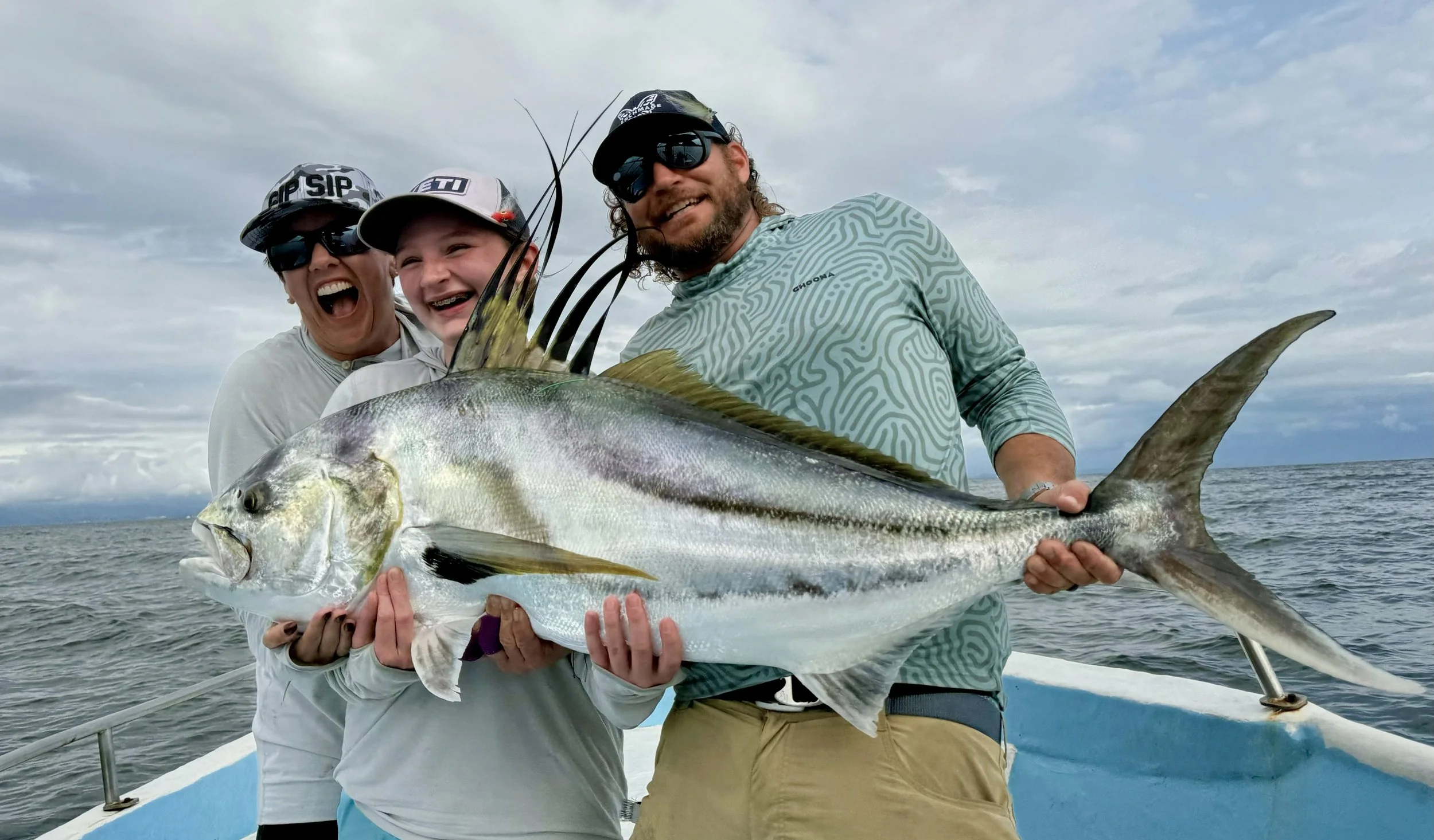 Drew Chicone, his wife, Susan, and daughter, Lucy, on a boat fishing crew holding a large rooster fish, smiling, with water and cloudy sky in the background.