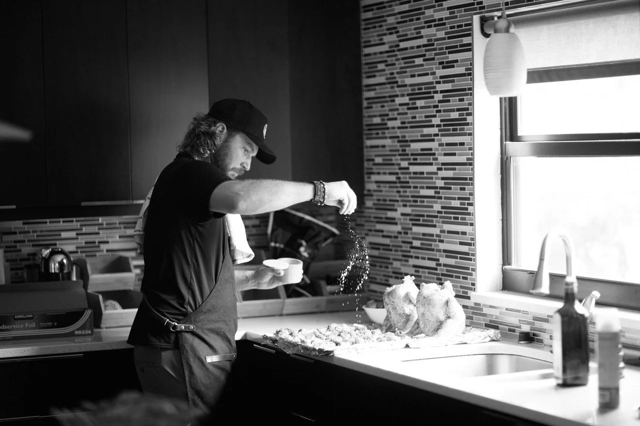 Drew Chicone cooking in his home kitchen, seasoning food on butchers block, with a window and tiled wall in the background.