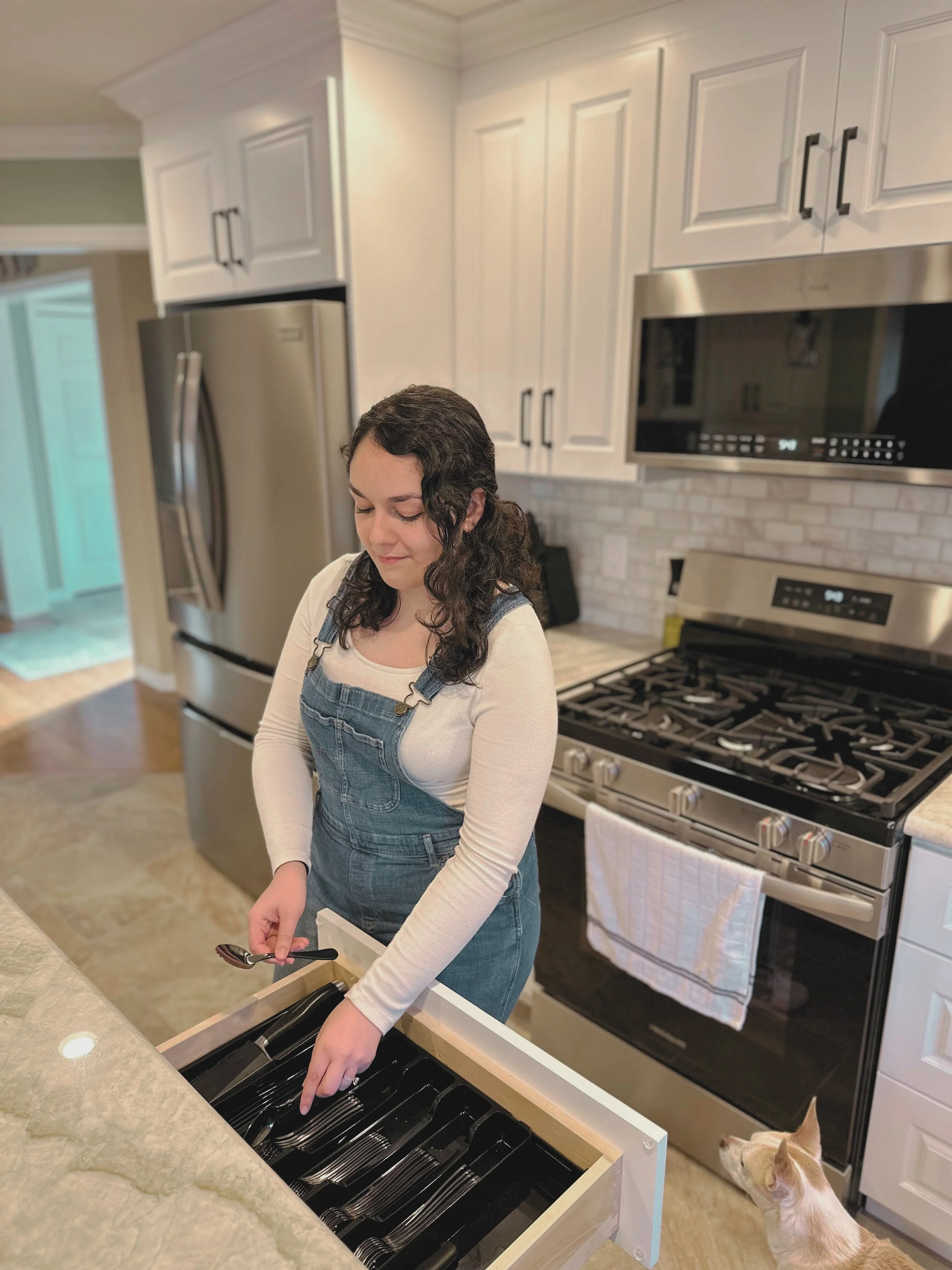 A woman loading silverware into a wooden drawer in a modern kitchen with white cabinets, a stainless steel refrigerator, and a gas stove. A cat looks up at her from the floor.