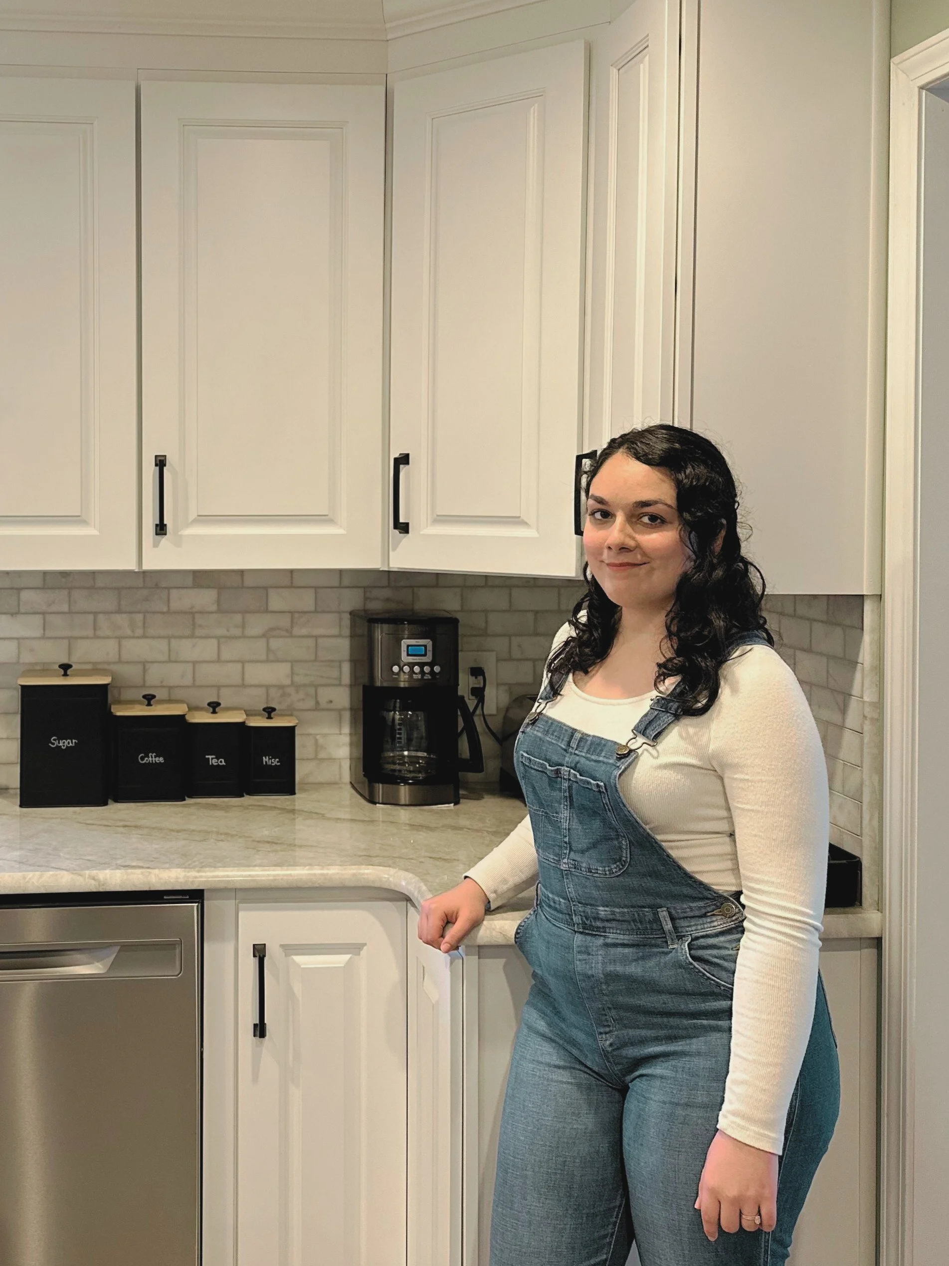A woman with curly dark hair, wearing a beige long-sleeve shirt and denim overalls, standing in a modern kitchen with white cabinets, a marble countertop, and black labeled canisters for sugar, coffee, tea, and miscellaneous items, along with a coffee maker.