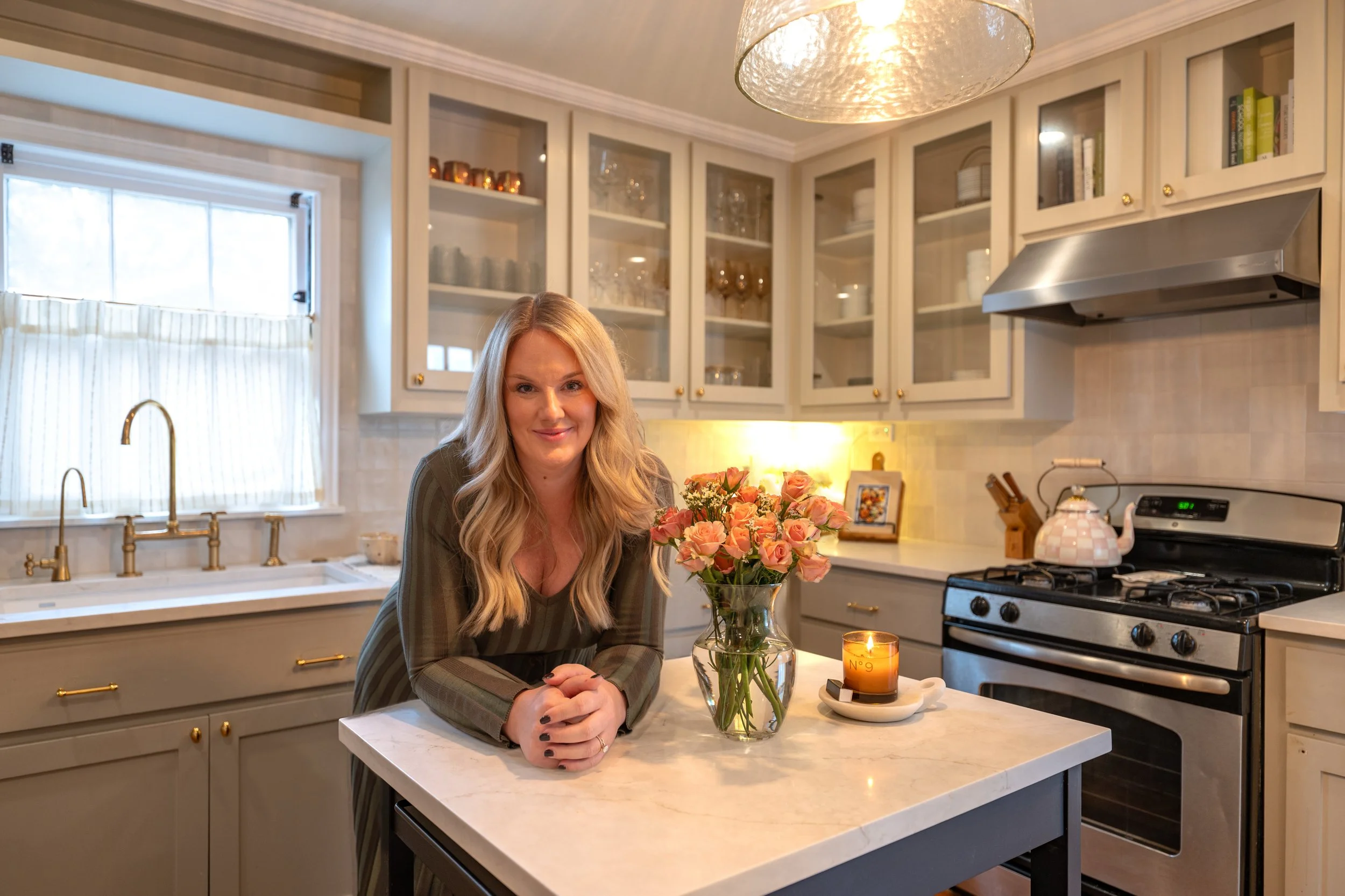 A woman with blonde hair smiling at the camera while leaning on a white kitchen island with a vase of pink roses and a lit candle. The kitchen has cream cabinets with glass doors, a window with white curtains, and stainless steel appliances.