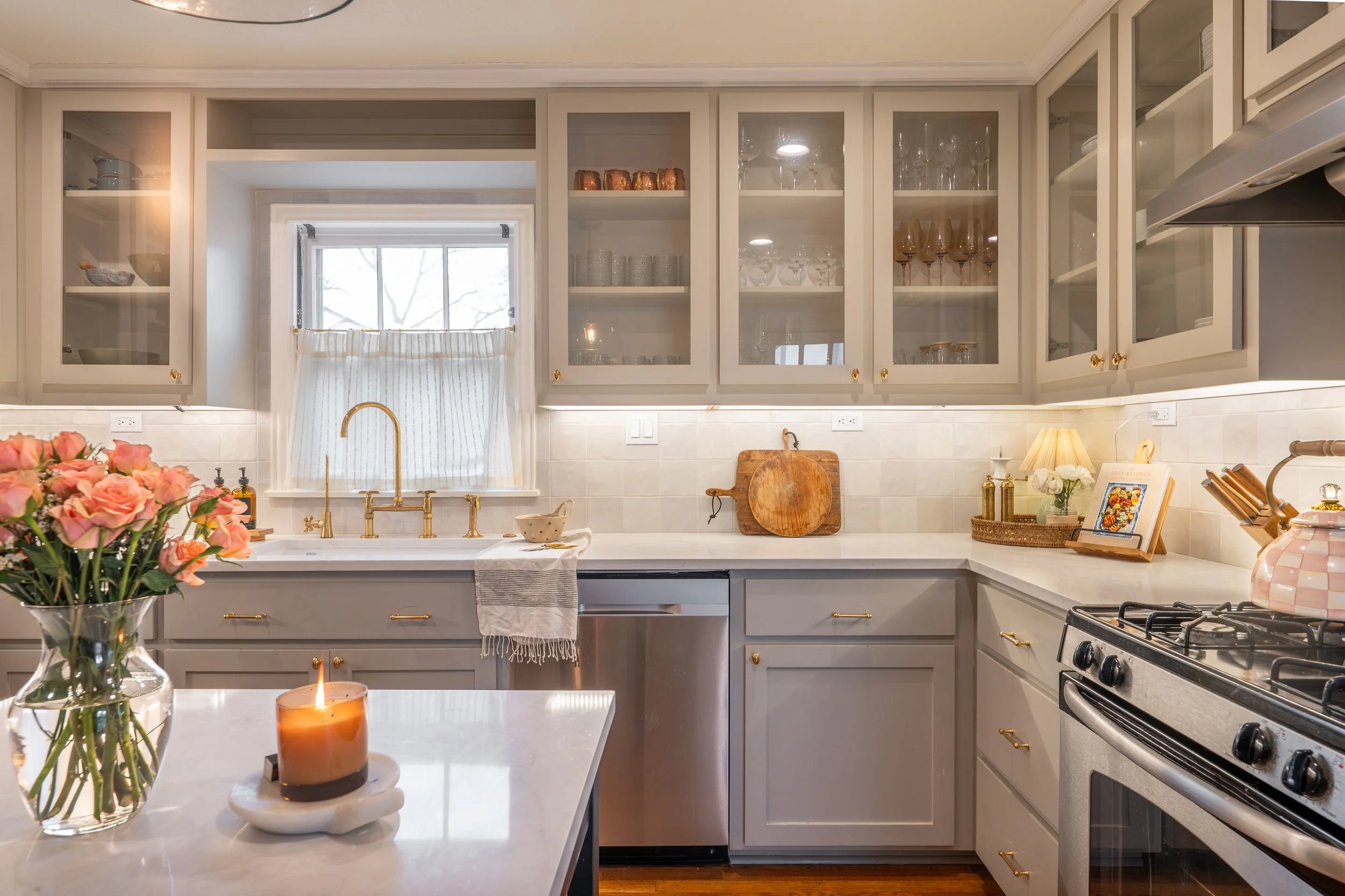 A cozy kitchen with white cabinets, a small window above the sink, and various kitchen items and decorations on the countertop, including flowers, a candle, and framed pictures.
