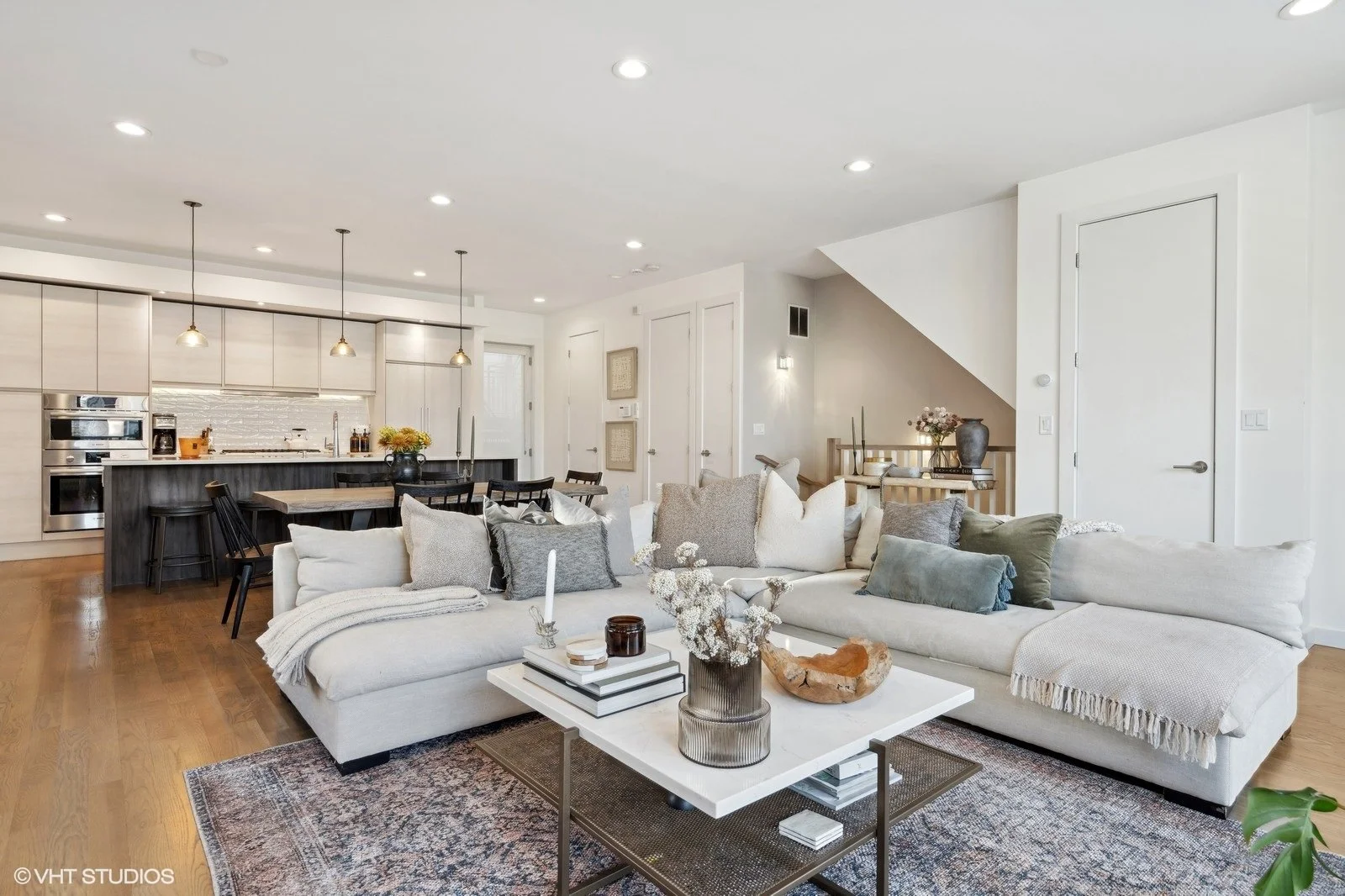Open-concept living room with white sectional sofa, coffee table, and a cozy throw blanket, connected to a modern kitchen with white cabinets, black island, and pendant lights.